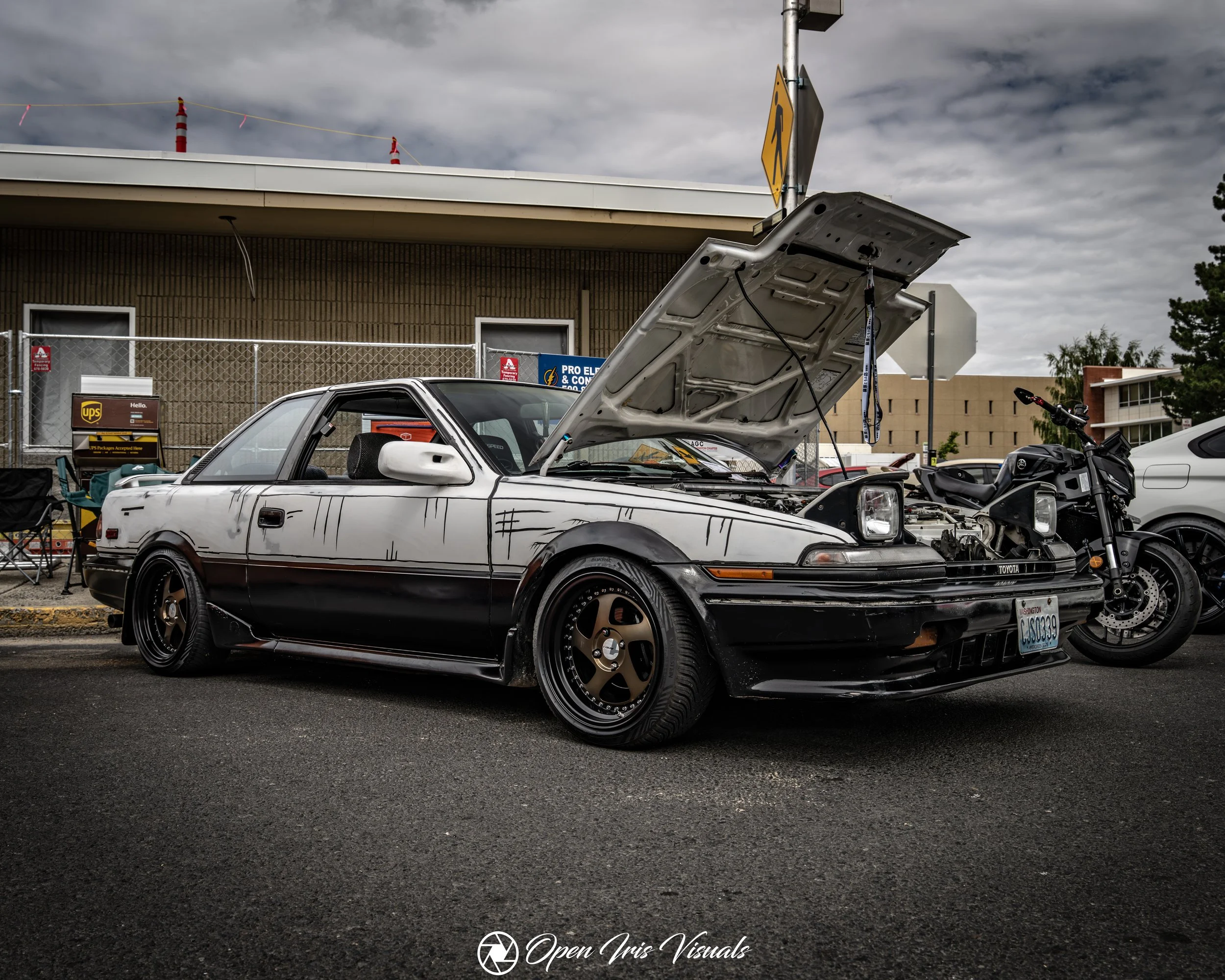 A modified two-door car with its hood open, parked on the street. It has a black and white paint job with graffiti-style streaks, bronze wheels, and a lowered stance. A motorcycle is parked next to it, and there are other vehicles and building structures in the background.