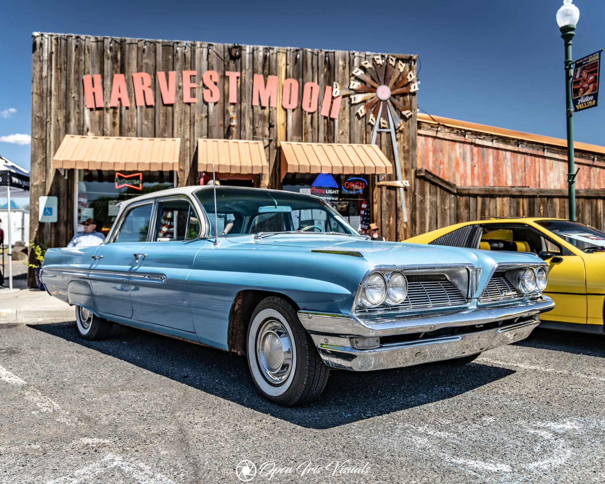 A vintage blue car parked in front of a wooden building with a sign that reads "Harvest Moon" and a windmill decoration. There are other cars in the parking lot, and a lamppost with a banner on it.