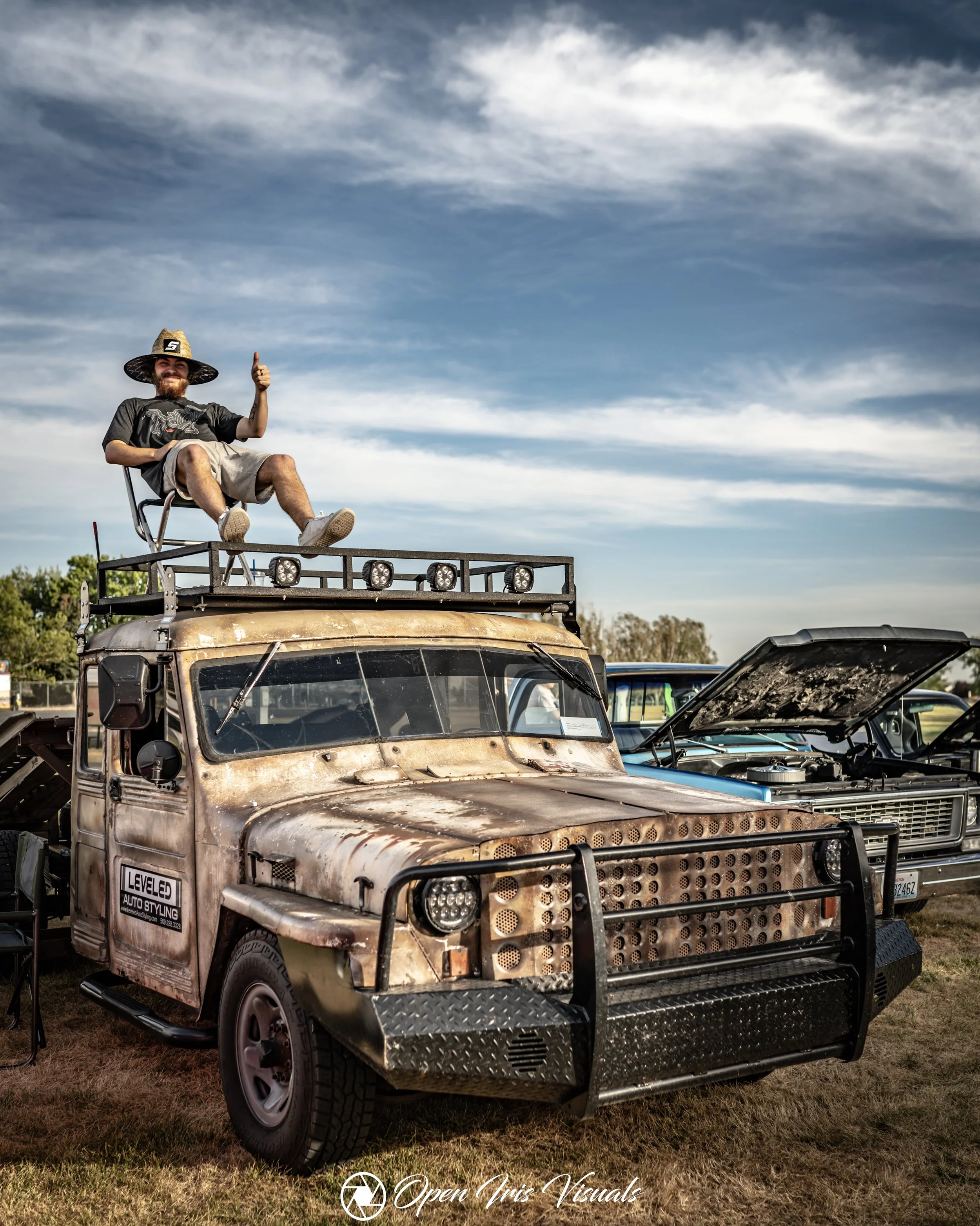 A man sitting on a chair on top of a rusty, vintage vehicle in an open field at an outdoor car show, giving a thumbs-up.