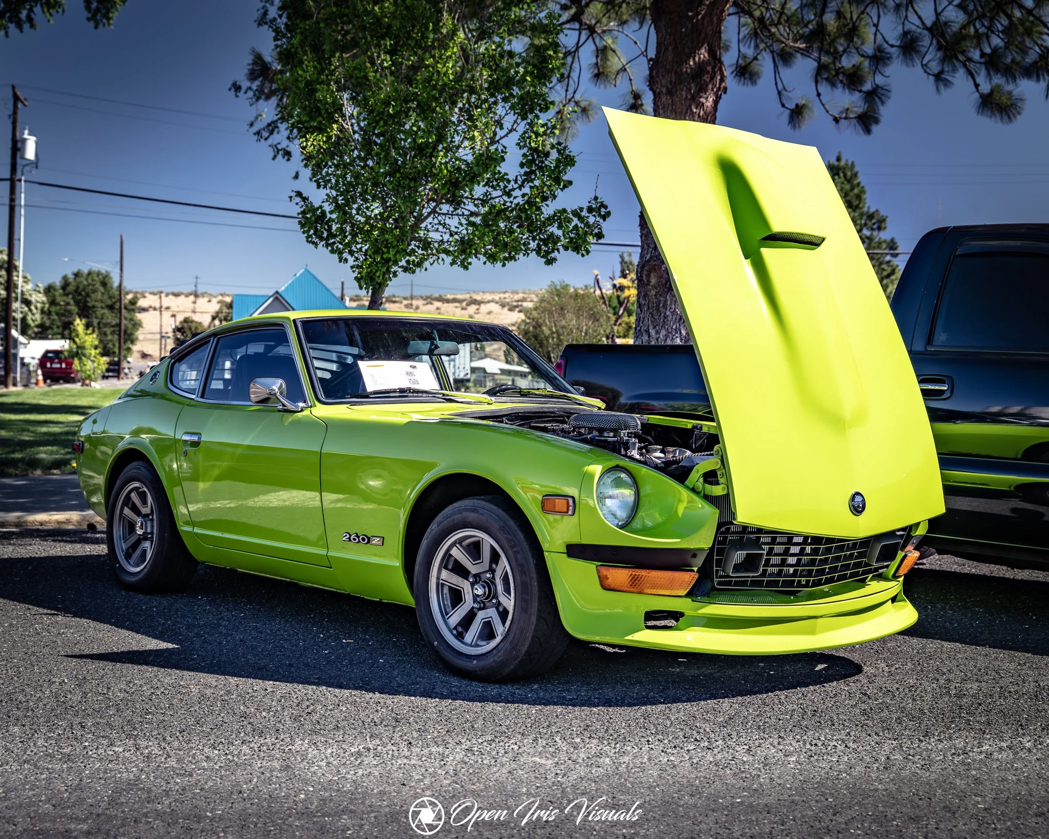A bright lime green vintage sports car with a sleek design and distinctive wheel rims, parked outdoors on a sunny day, with its hood open revealing the engine. There are trees and utility poles in the background.