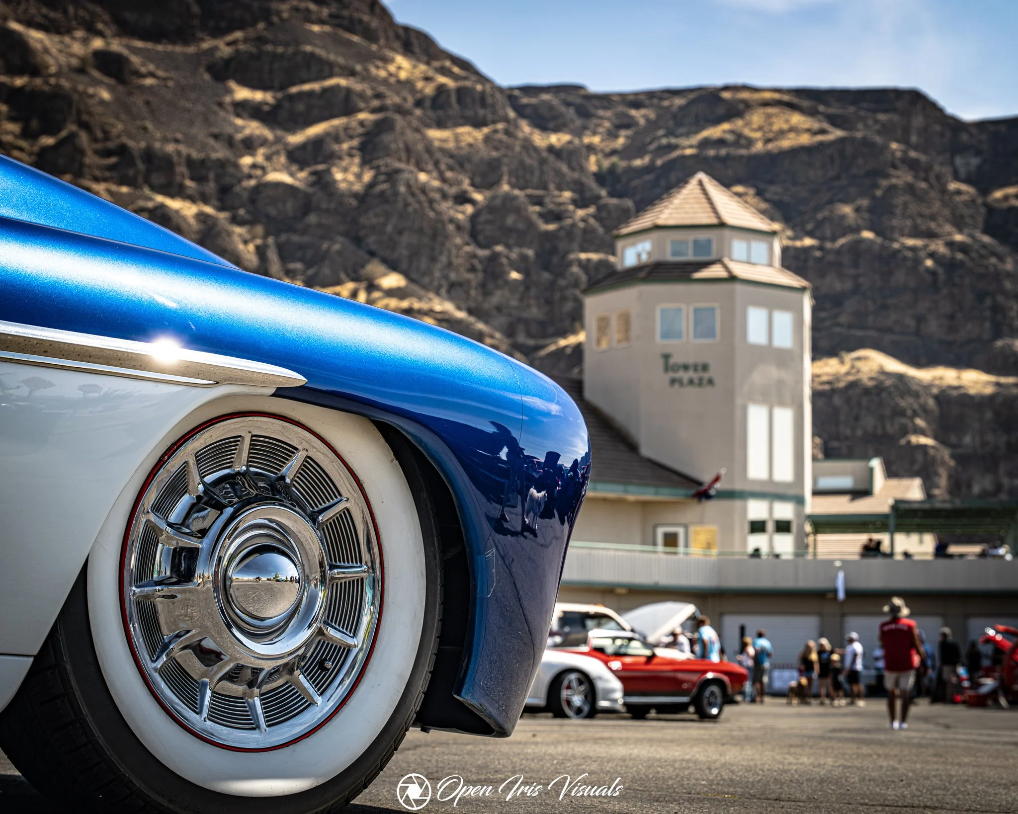 Close-up of a vintage blue and white car's front wheel, with a shiny chrome hubcap, at a car show. In the background, there are several other cars, a building labeled 'Tower Plaza,' and a rocky hillside under a clear sky.