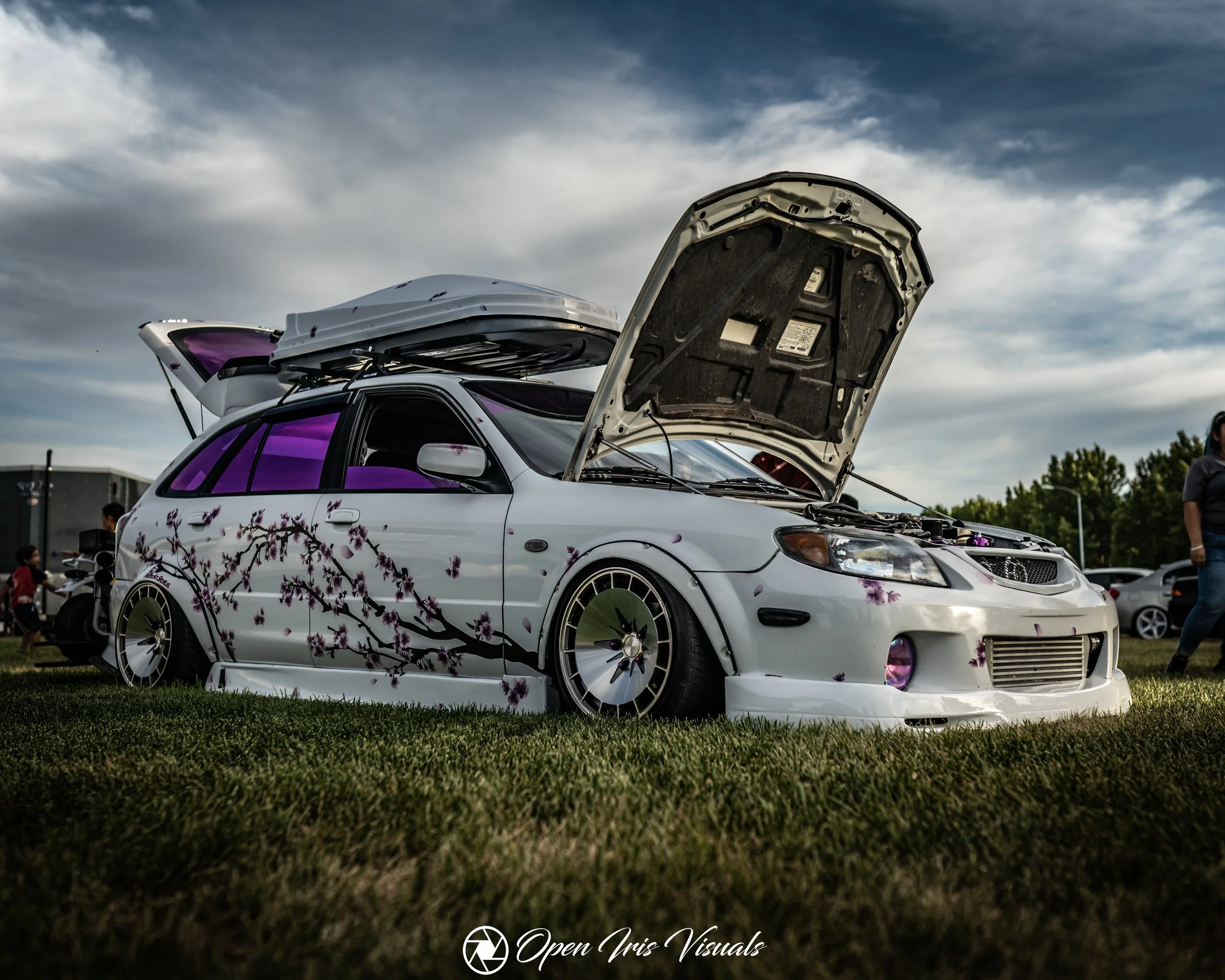 White modified car with cherry blossom decals, lowered stance, purple interior, and open hood at a car show on grass under cloudy sky.