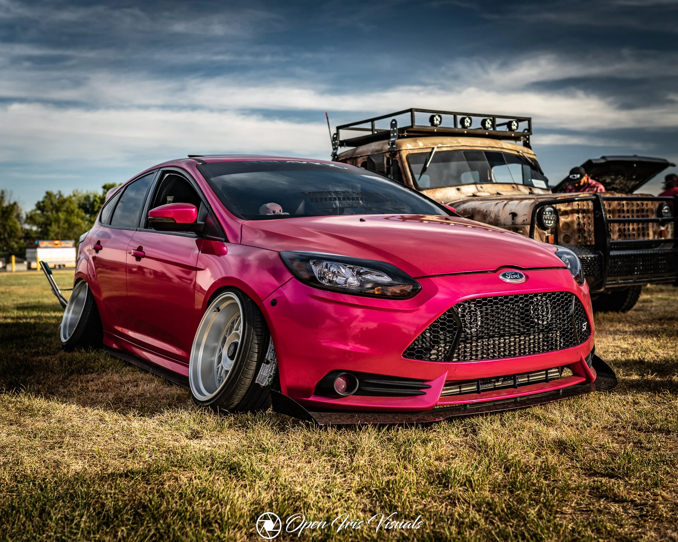 A heavily modified pink Ford Focus with lowered suspension and custom wheels parked on grass at a car show, with an old, rusted truck in the background under a partly cloudy sky.