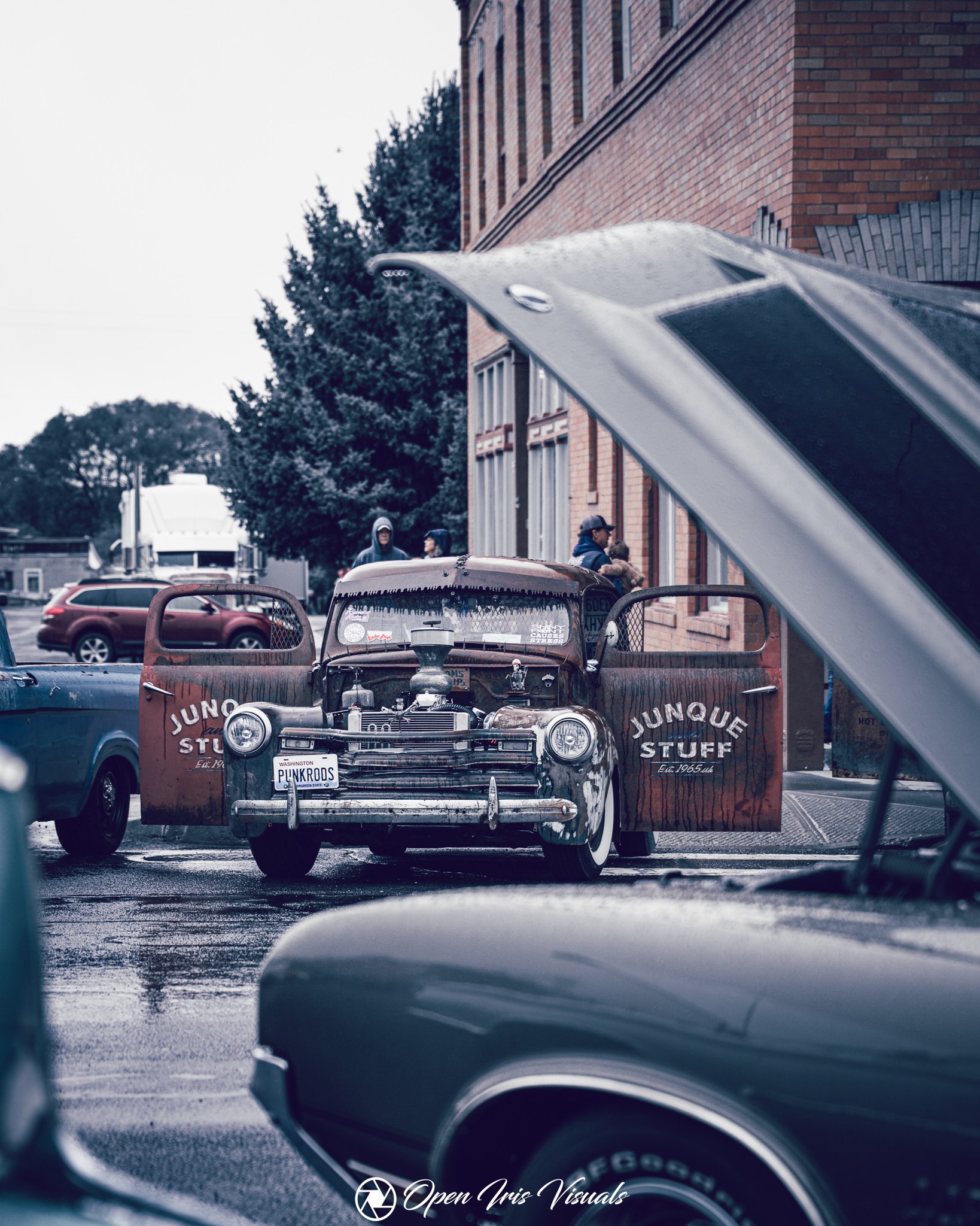 View through a car's interior shows a worn pickup truck with a opened hood in front of a brick building, with people standing and trees in the background on an overcast day.