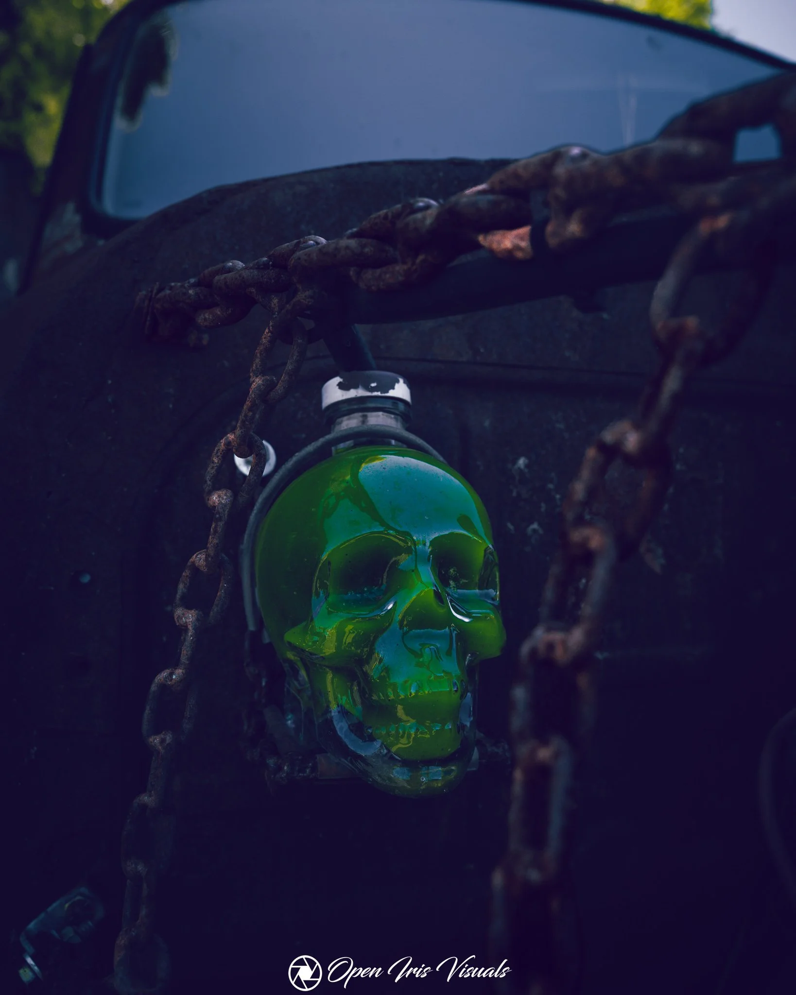 A green skull sculpture with reflections, placed underneath a rusted chain and a bottle of water, against a dark background with a faint outdoor setting at the top.