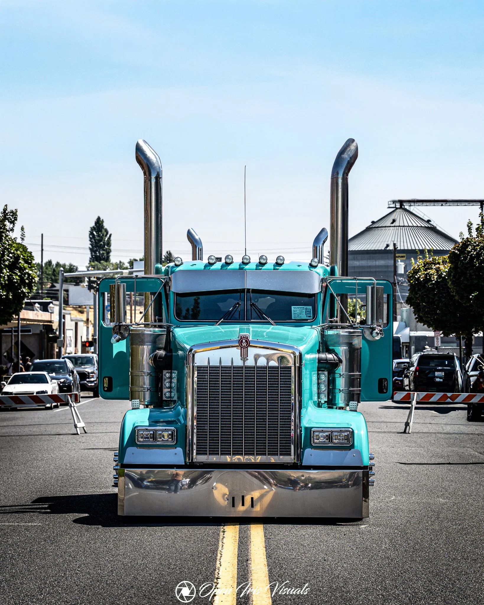 A turquoise semi-truck with chrome exhaust stacks and a large front grill parked in a parking lot, with a grain silo and trees in the background under a clear blue sky.