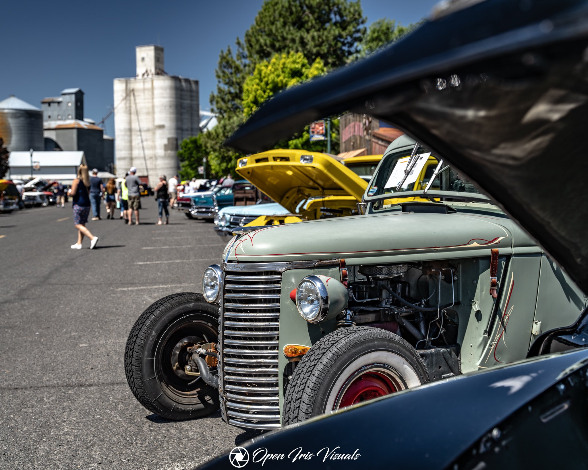 A vintage car with its hood open is displayed at a car show, with other classic cars and attendees in the background, and silos and trees on a sunny day.