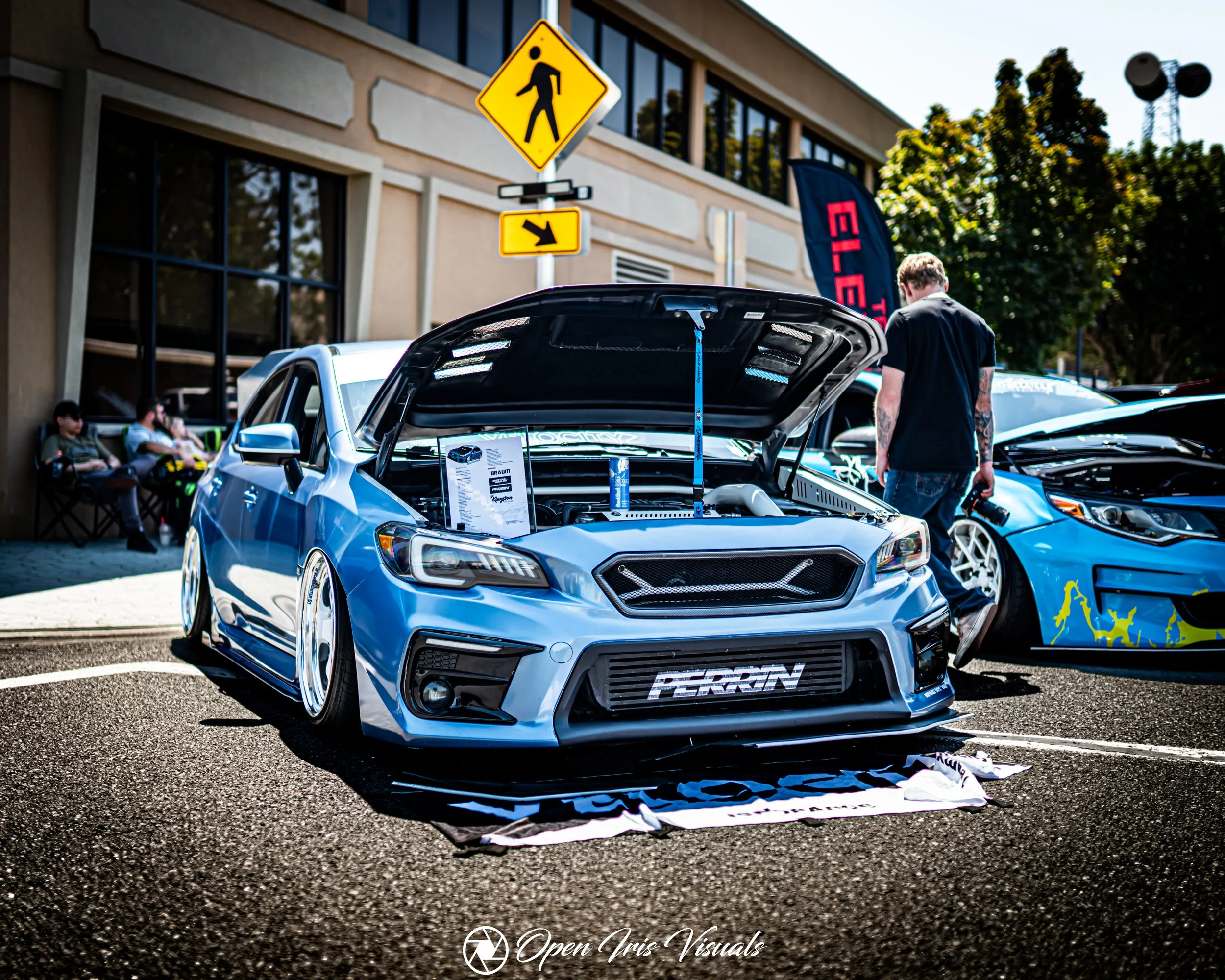 A blue modified car with its hood open at a car show, displaying custom parts, with a person looking at it and other cars and spectators in the background.