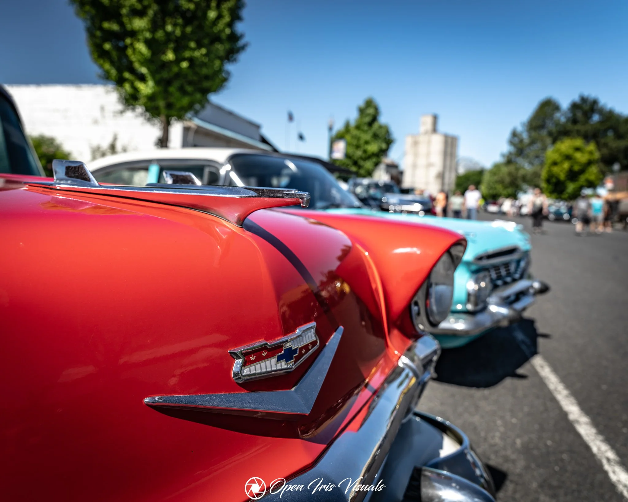 Close-up of a red vintage Chevrolet car showing the Chevrolet emblem on the front fender. In the background, there are other classic cars parked, trees, and a building under a clear blue sky.