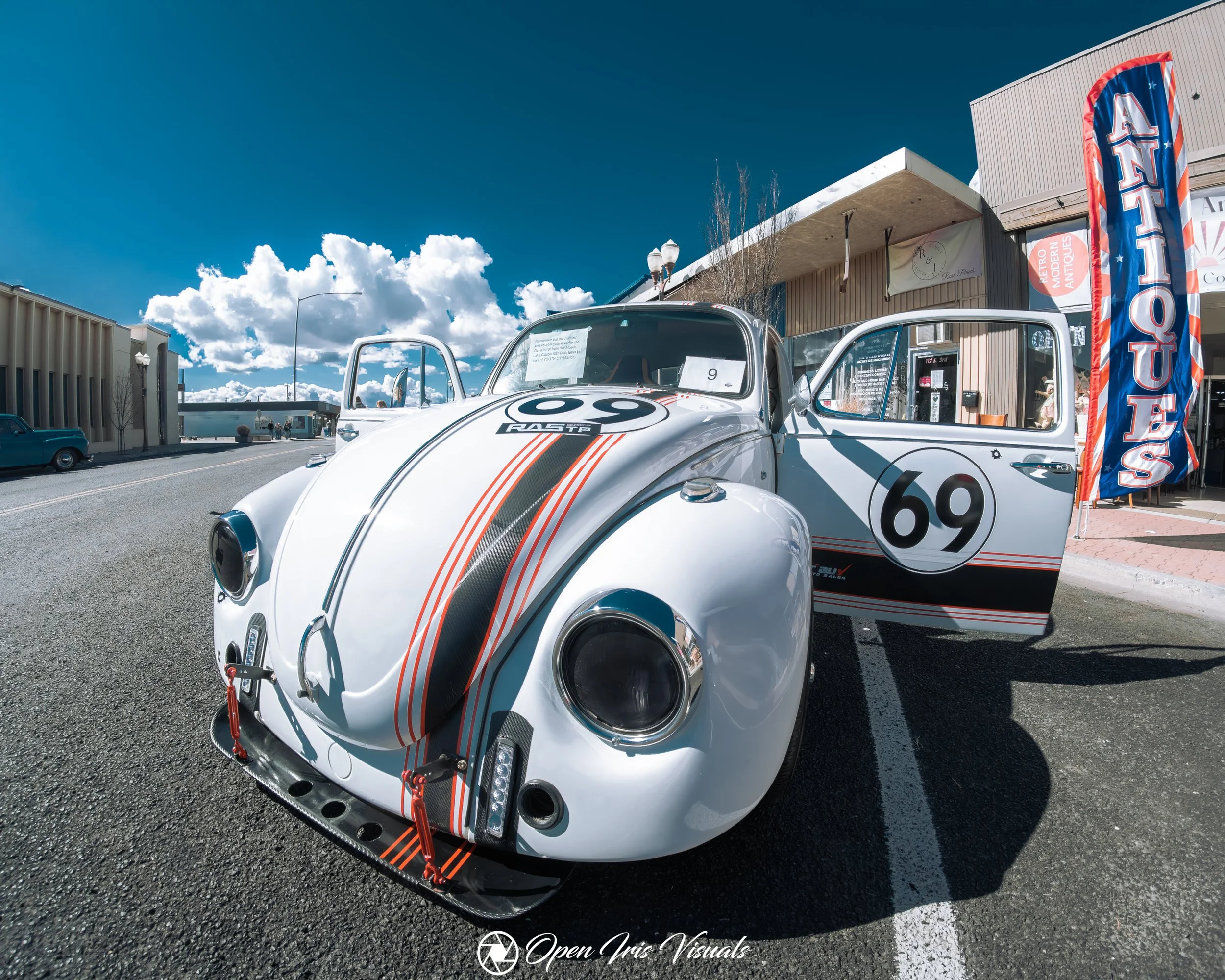 A vintage VW Beetle race car with the number 69 on the side and front, featuring a white body with black and orange racing stripes, parked outside a building with a banner that reads 'ANTIQUE.'