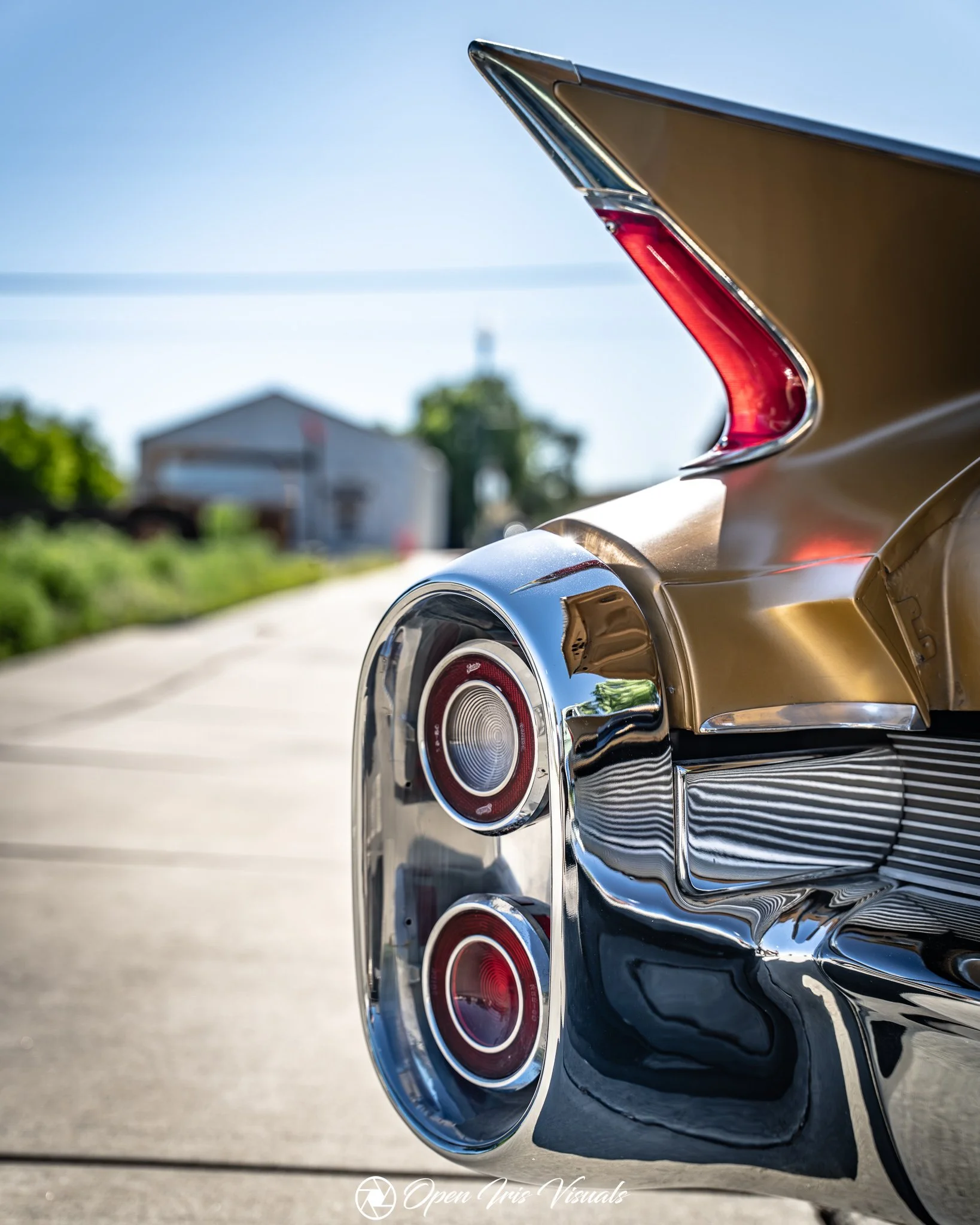 Close-up of the rear tail lights and bumper of a vintage gold and chrome car, with a blurred house and trees in the background.