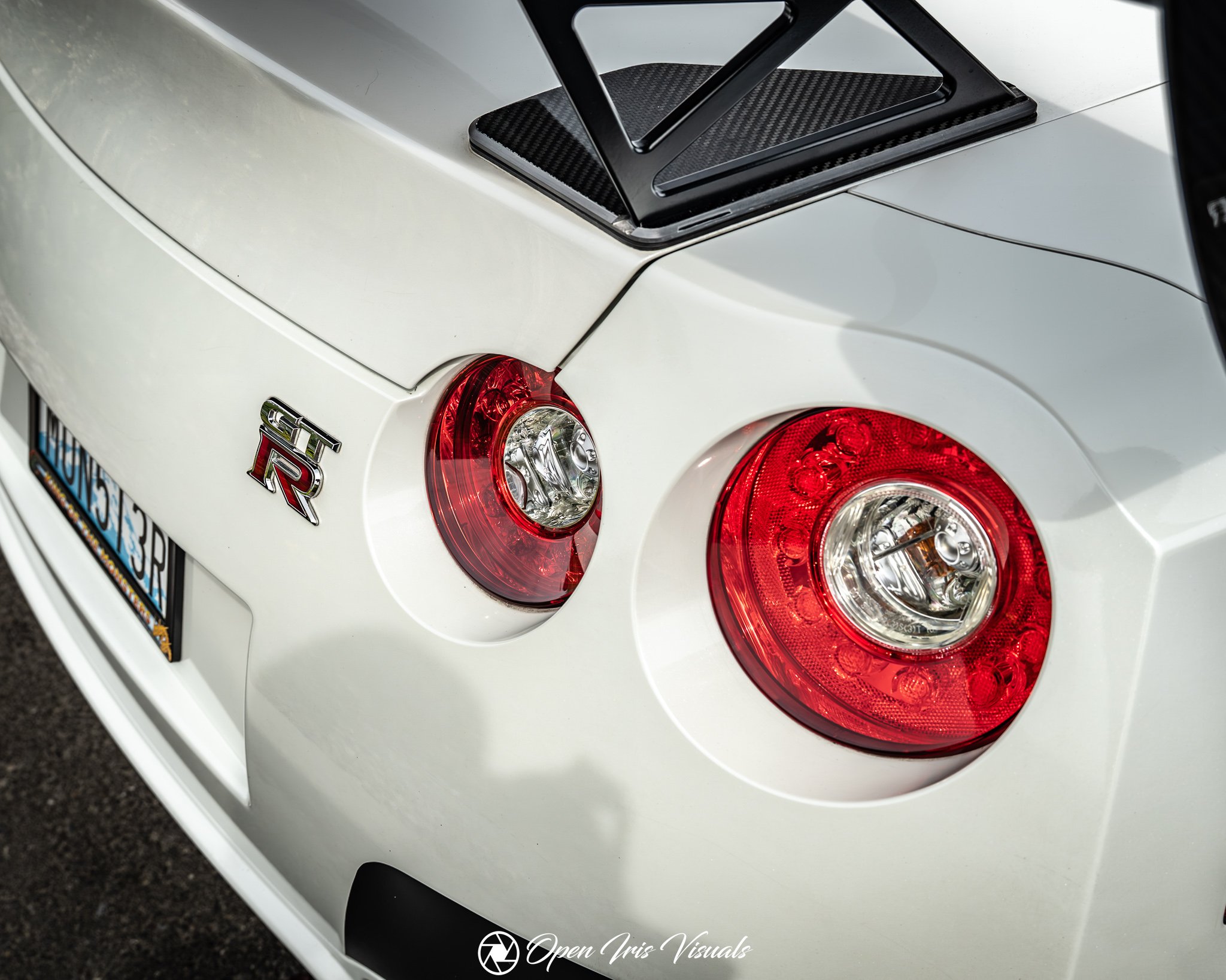 Close-up of a white Nissan Skyline GTR's rear end, showing red and clear taillights, a black carbon fiber rear wing, and an emblem with the letters 'GT-R' underneath.