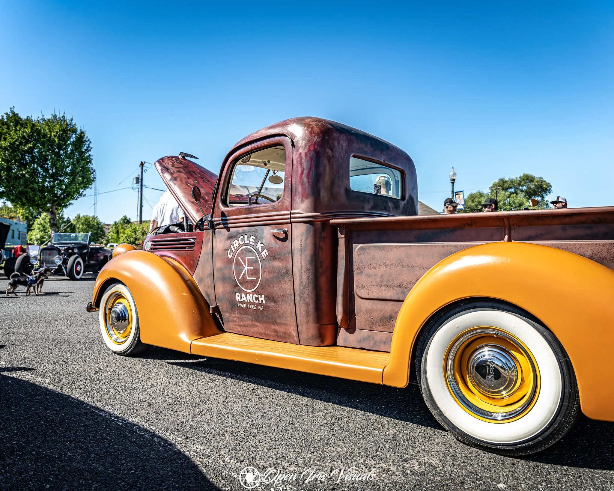Vintage rusty truck with yellow fenders parked at a car show, with other classic cars and people in the background under a clear blue sky.
