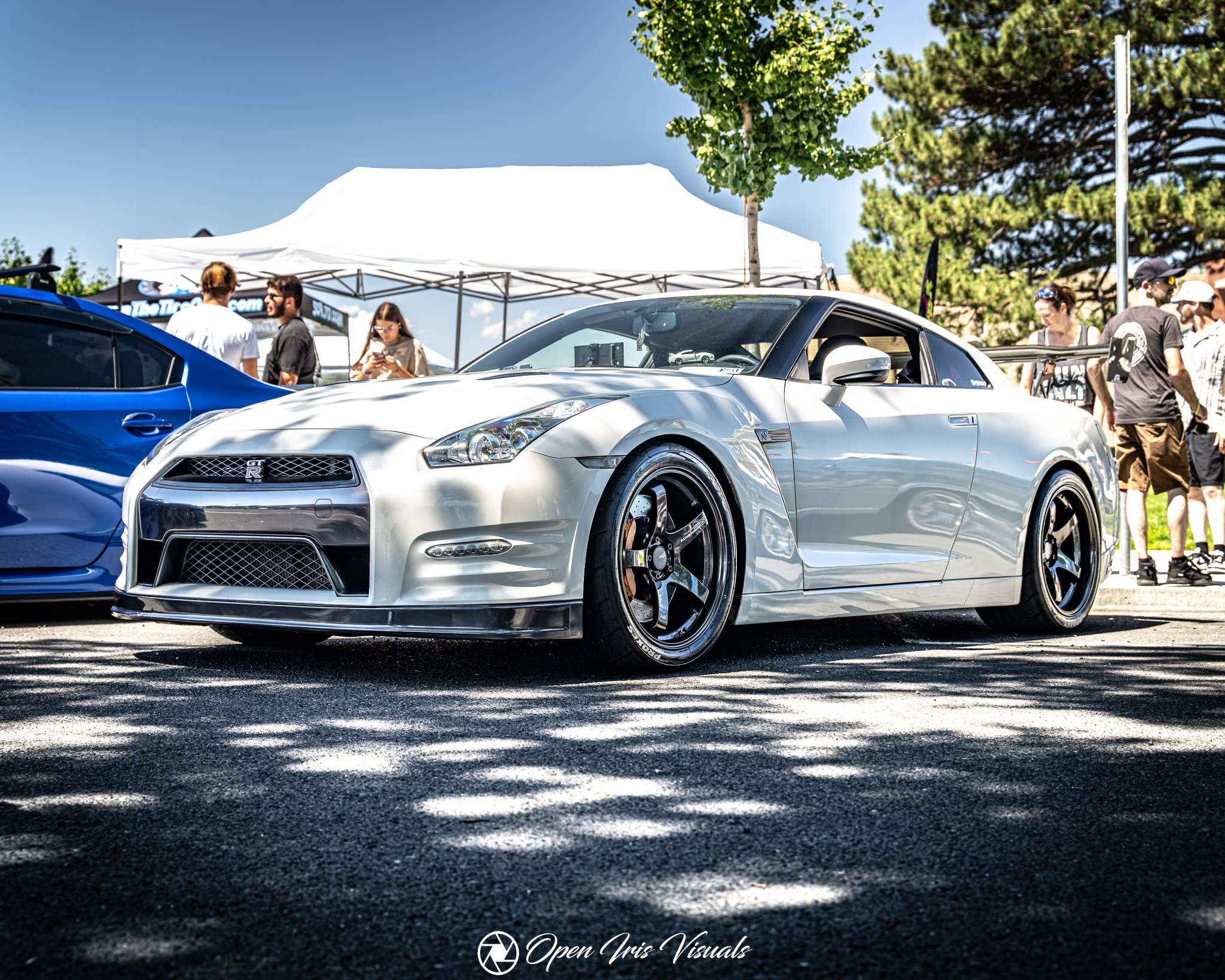 A silver Nissan GT-R sports car parked outdoors at a car show, surrounded by people and tents, with trees and a clear blue sky in the background.