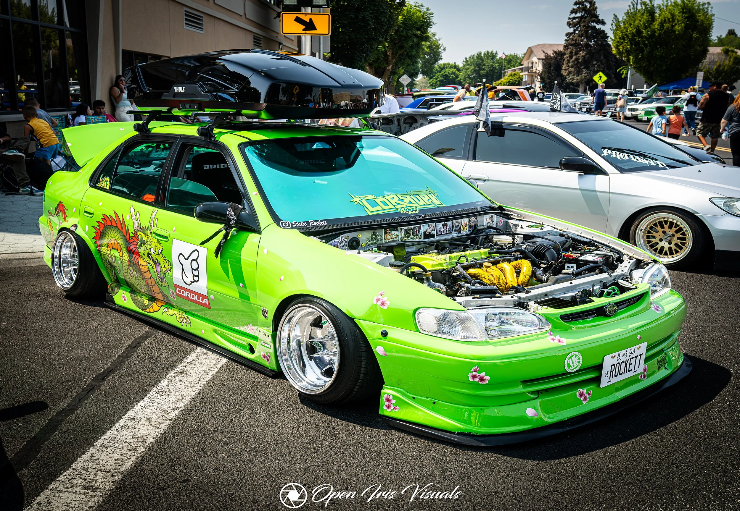 A vibrant lime green car decorated with floral designs and a dragon decal, with its hood open exposing the engine, parked among other cars at a car show. There are people walking and sitting nearby, and a building with a yellow arrow sign above.