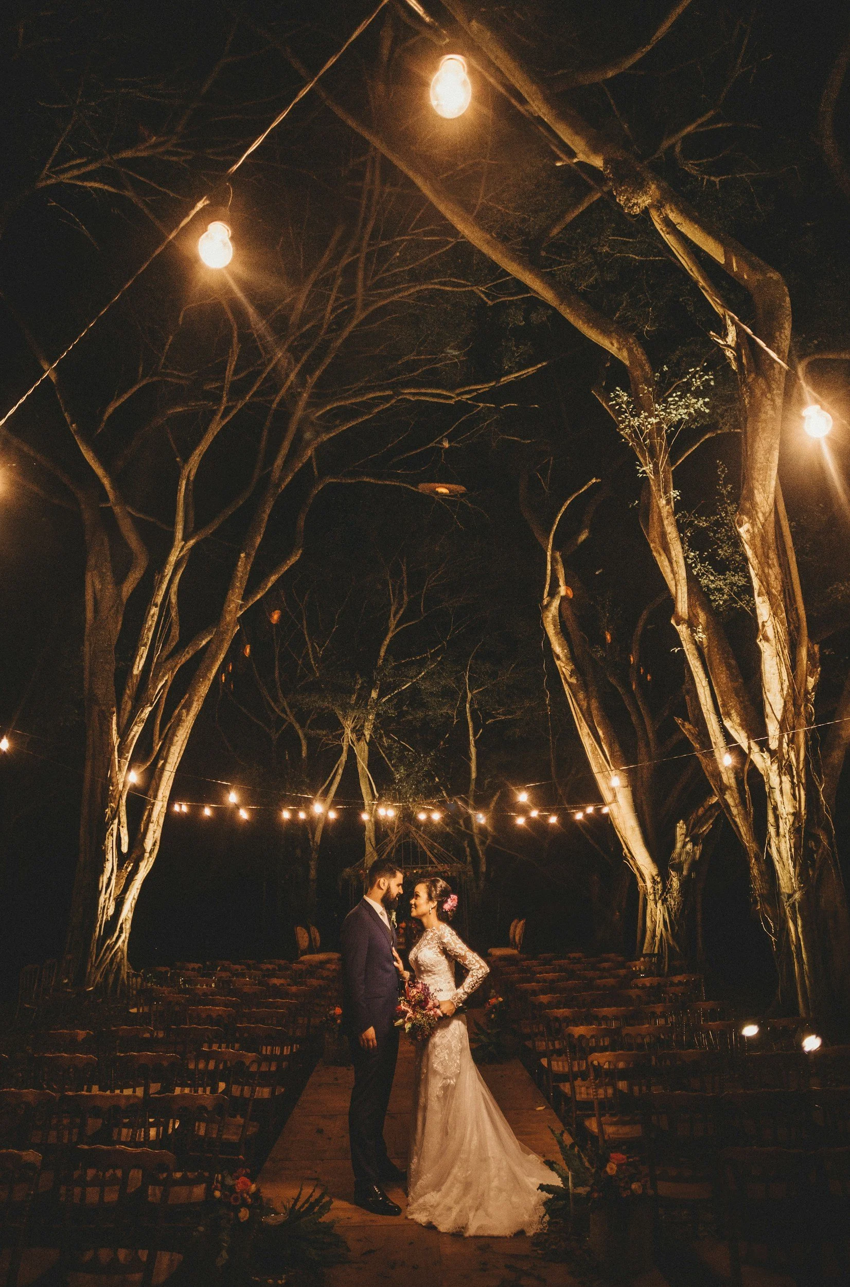 Pareja de novios en una boda en la noche, entre árboles iluminados con luces colgantes, en un entorno rústico y romántico.