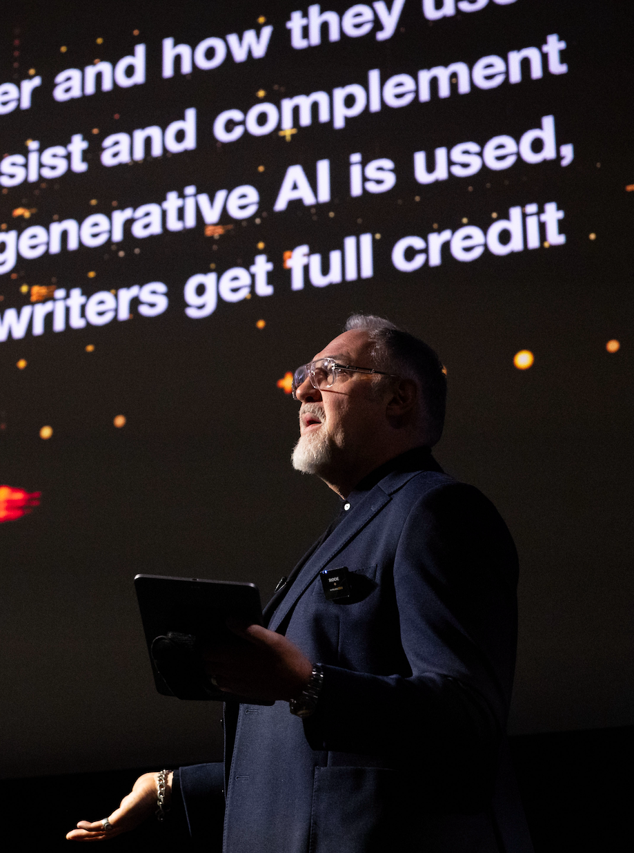 A man in a dark suit and glasses giving a presentation, holding a tablet, with a large screen behind him displaying partially visible text about AI and writers.