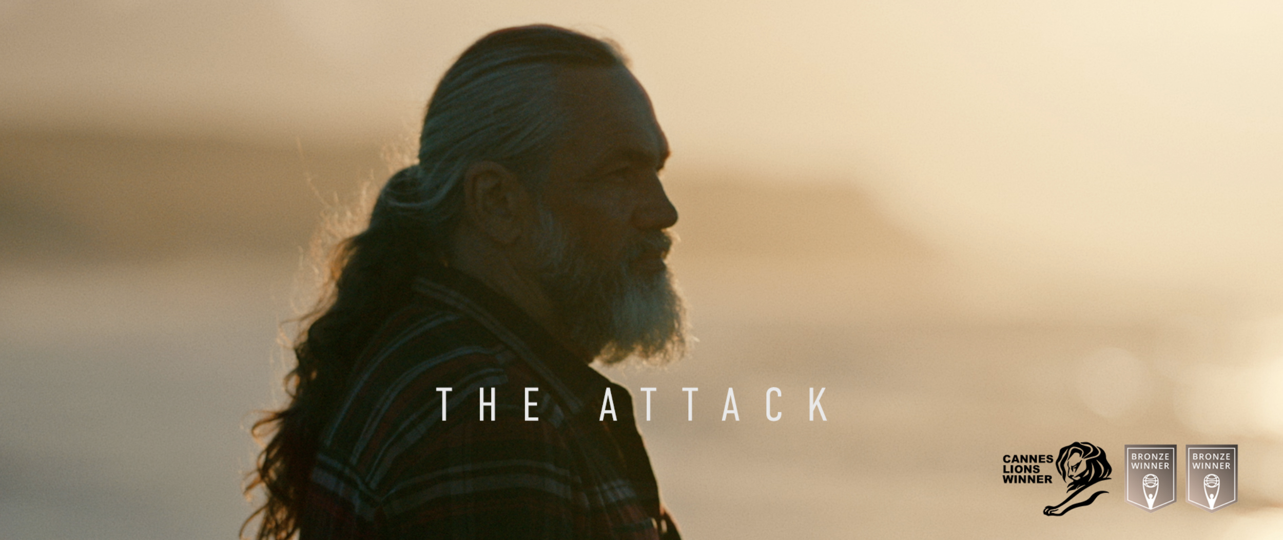 Side profile of an older man with long hair and a beard, looking toward the horizon during sunset, with the text 'The Attack' and awards in the bottom right corner.