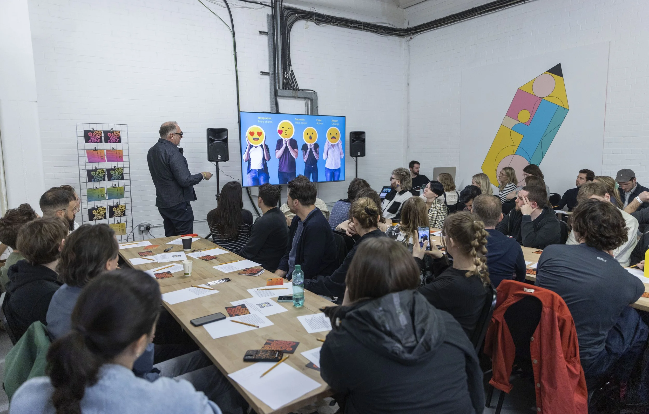 A large group of people attending a presentation or conference in a spacious room with white brick walls. The presenter, a man with gray hair and glasses, stands at the front near a large screen displaying four emojis depicting happiness, sadness, fear, and anger. Many attendees are seated at long tables with papers, notebooks, pens, and drinks in front of them.