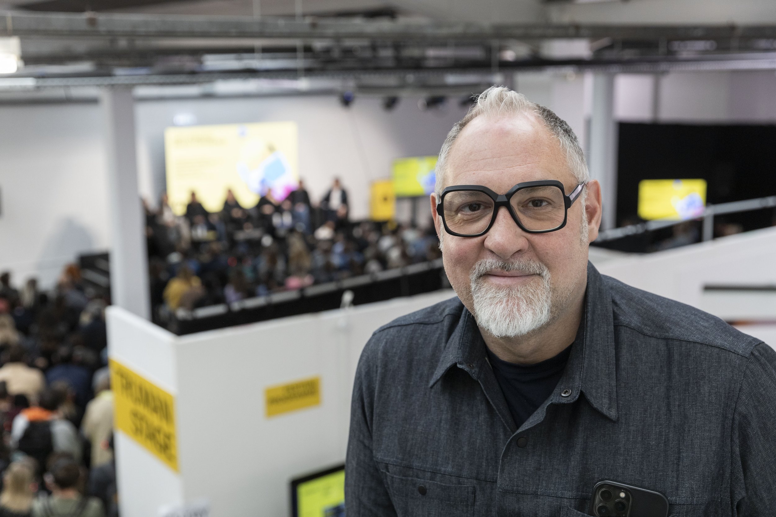 A man with glasses and a beard, wearing a dark shirt, taking a selfie at a crowded indoor event or conference.
