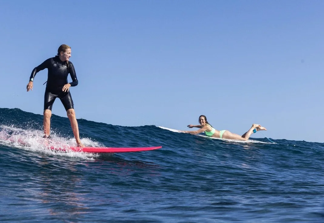 A mom rides a wave while her daughter paddles out smiling in Fiji