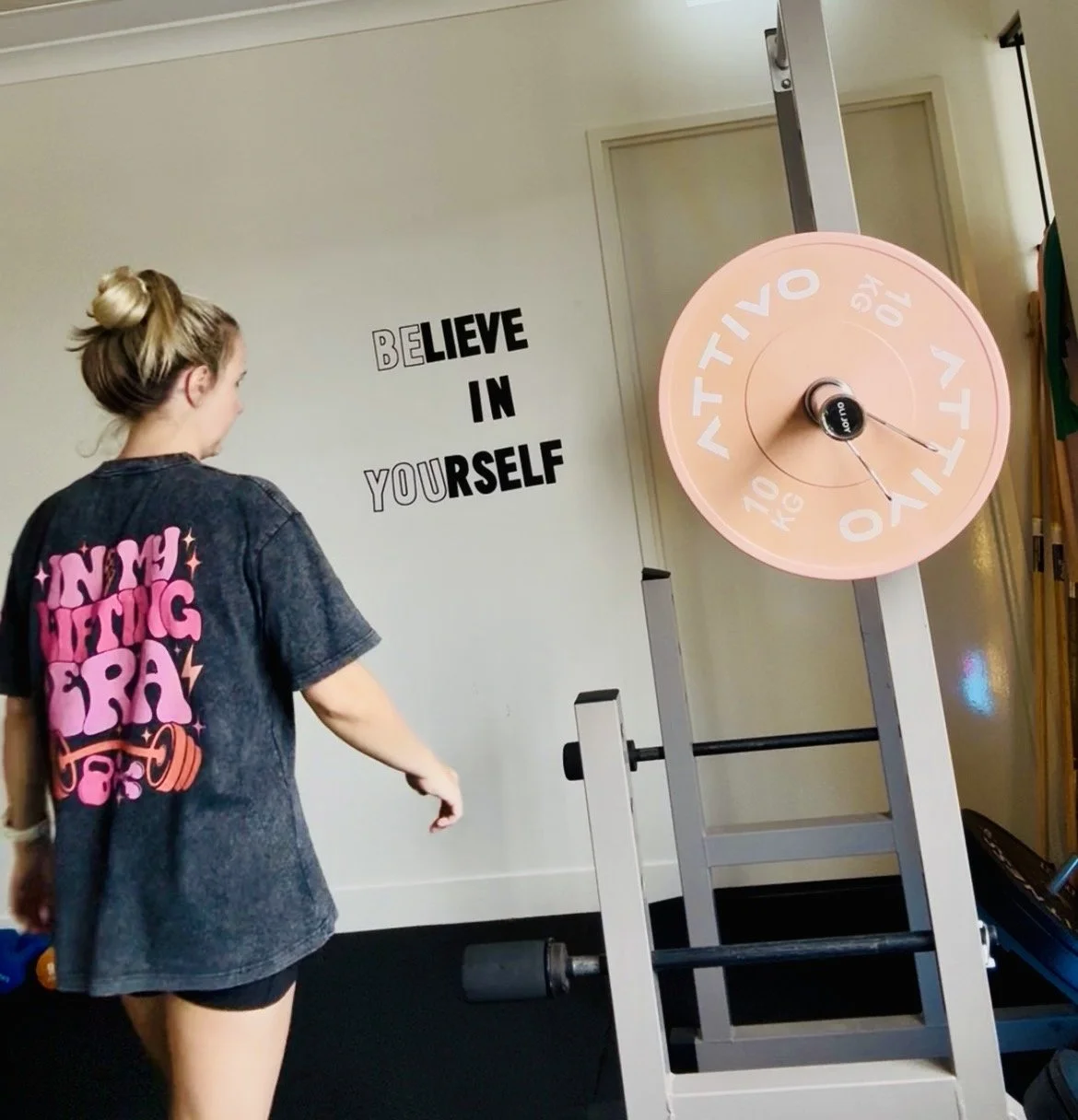 A woman with blonde hair in a bun, wearing a black t-shirt with pink and orange text, pointing at a pink weight plate on a squat rack in a gym. The wall behind her has motivational text that reads 'BELIEVE IN YOURSELF' in black letters.