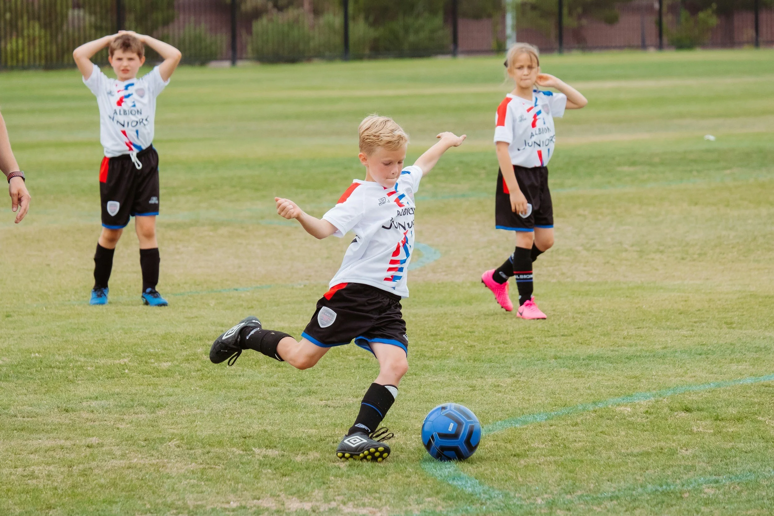 Young boys and girls playing soccer on a grassy field, with a boy kicking a blue soccer ball in the foreground and other children watching in the background.