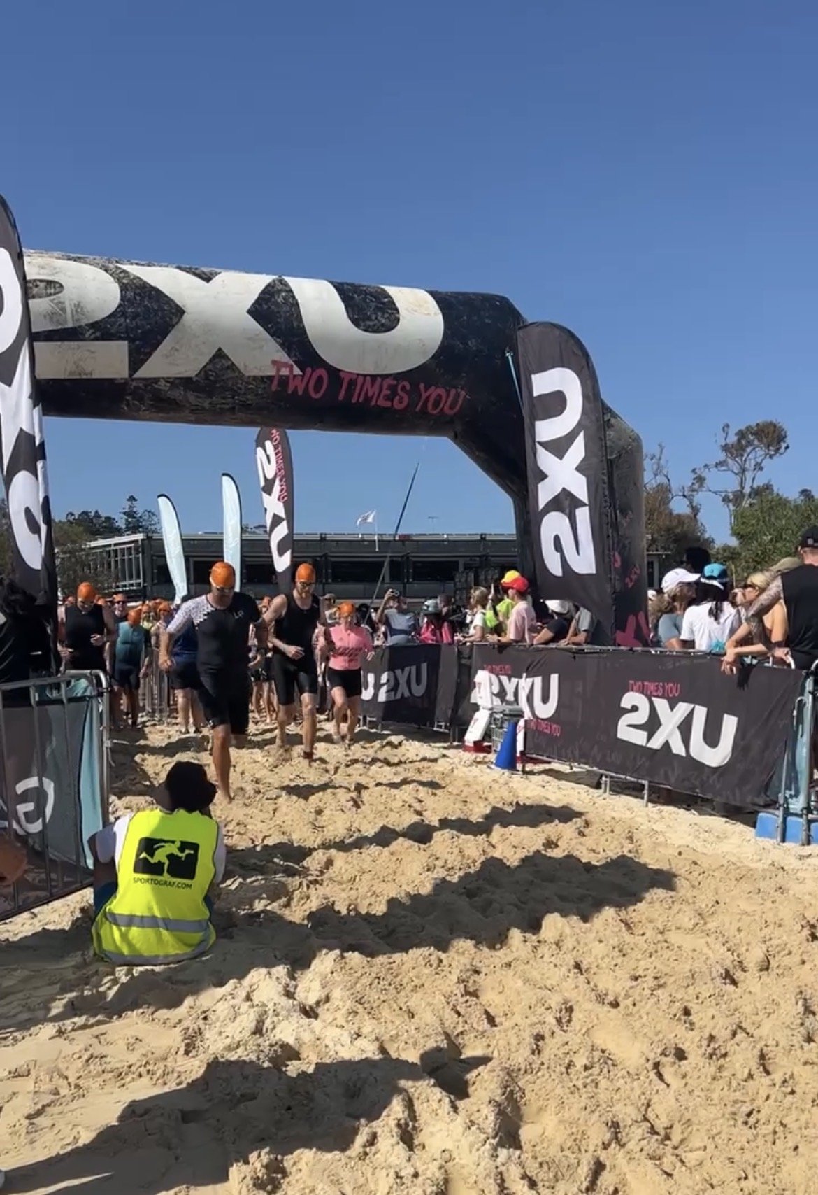 Participants start a race or triathlon event on a sandy beach, with a large black inflatable arch that has the brand name '2XU' and the slogan 'TWO TIMES YOU'. Spectators and event staff watch as runners in black suits and orange swim caps enter the water, indicating the beginning of the race.