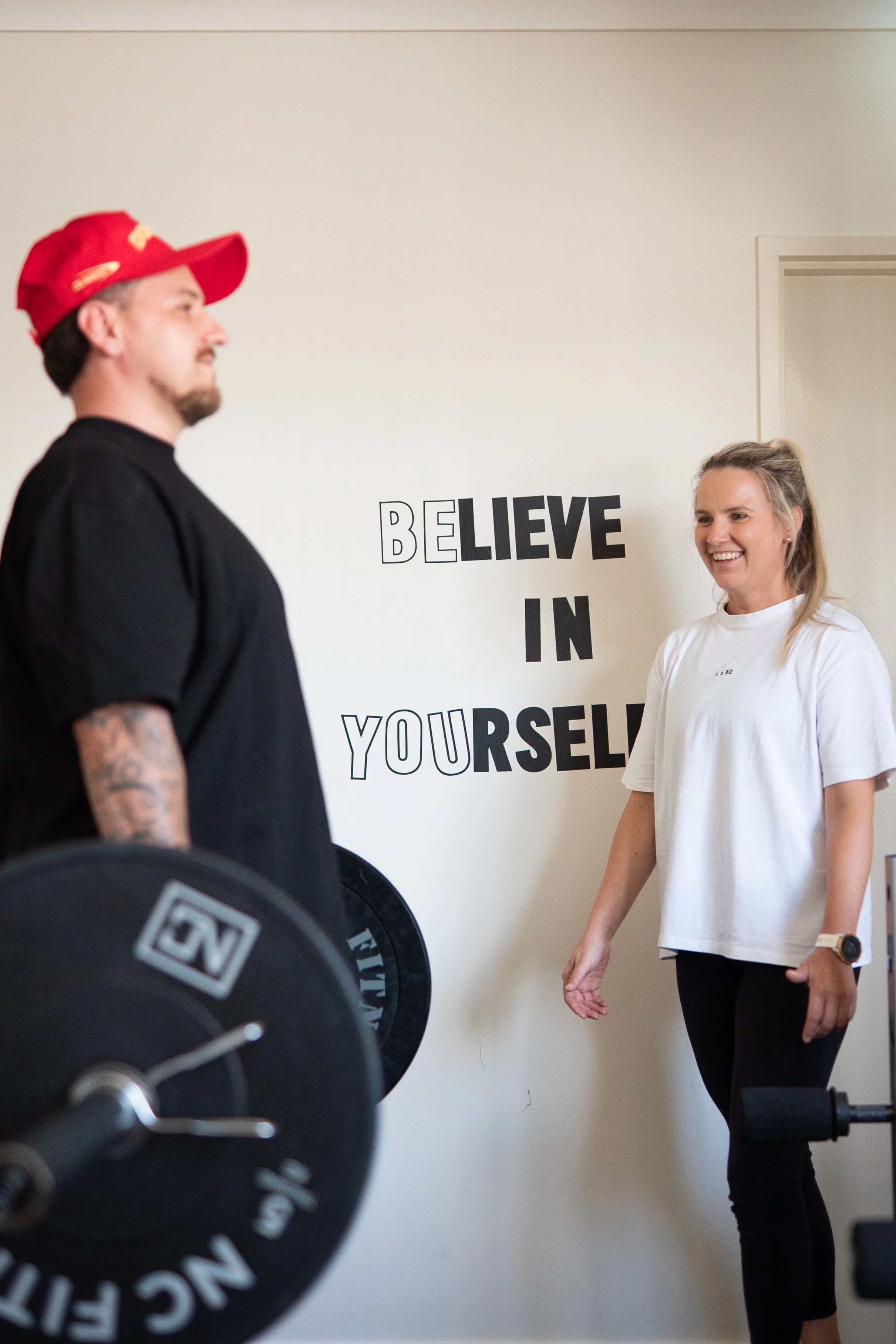 A man lifting weights in a gym while a woman smiles at him. The wall behind them has a motivational quote that reads 'BELIEVE IN YOURSELF'.