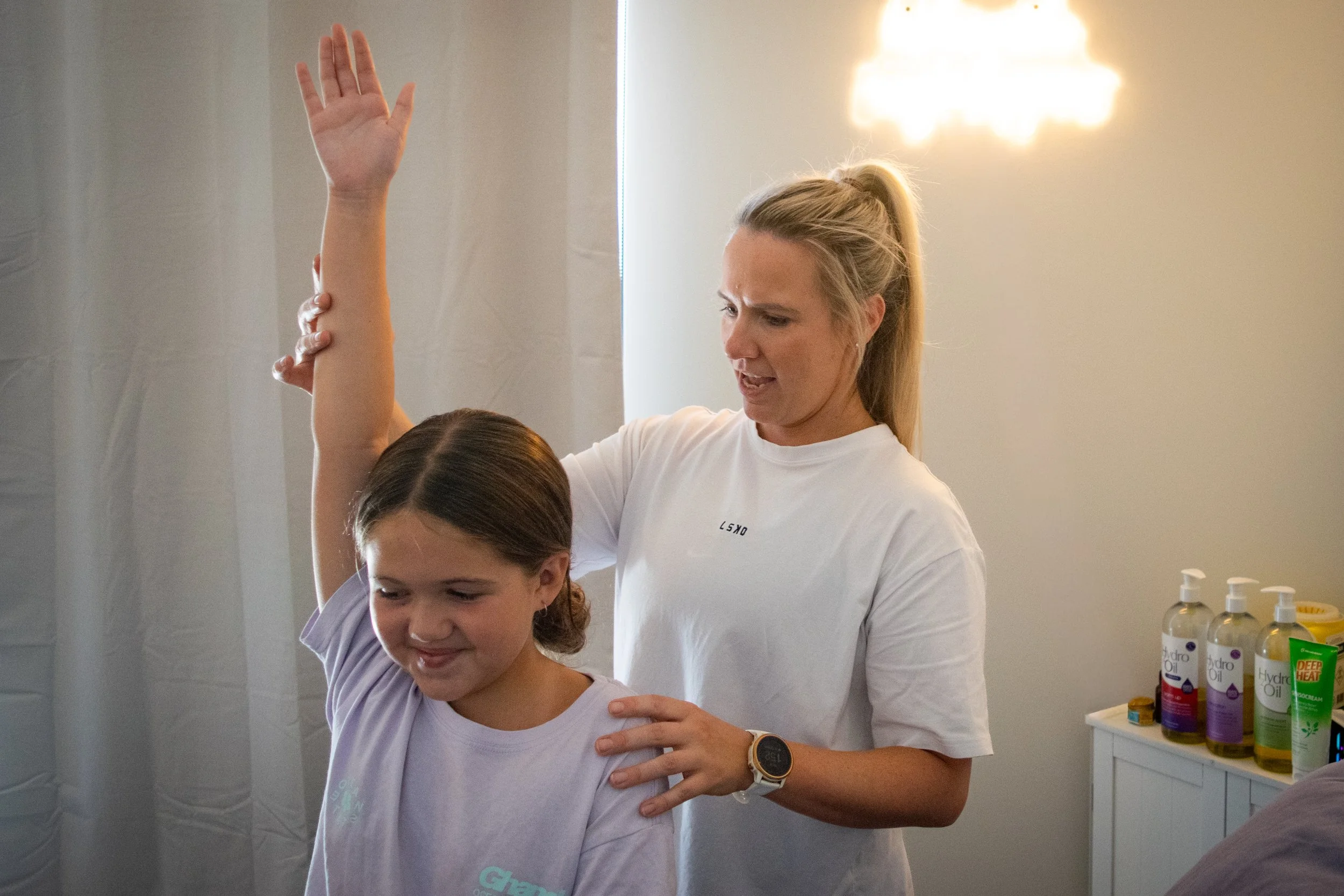 A woman assisting a young girl with a physical therapy exercise, the girl has her arm raised up as the woman supports her. The background shows a lamp and bottles of oil on a shelf.