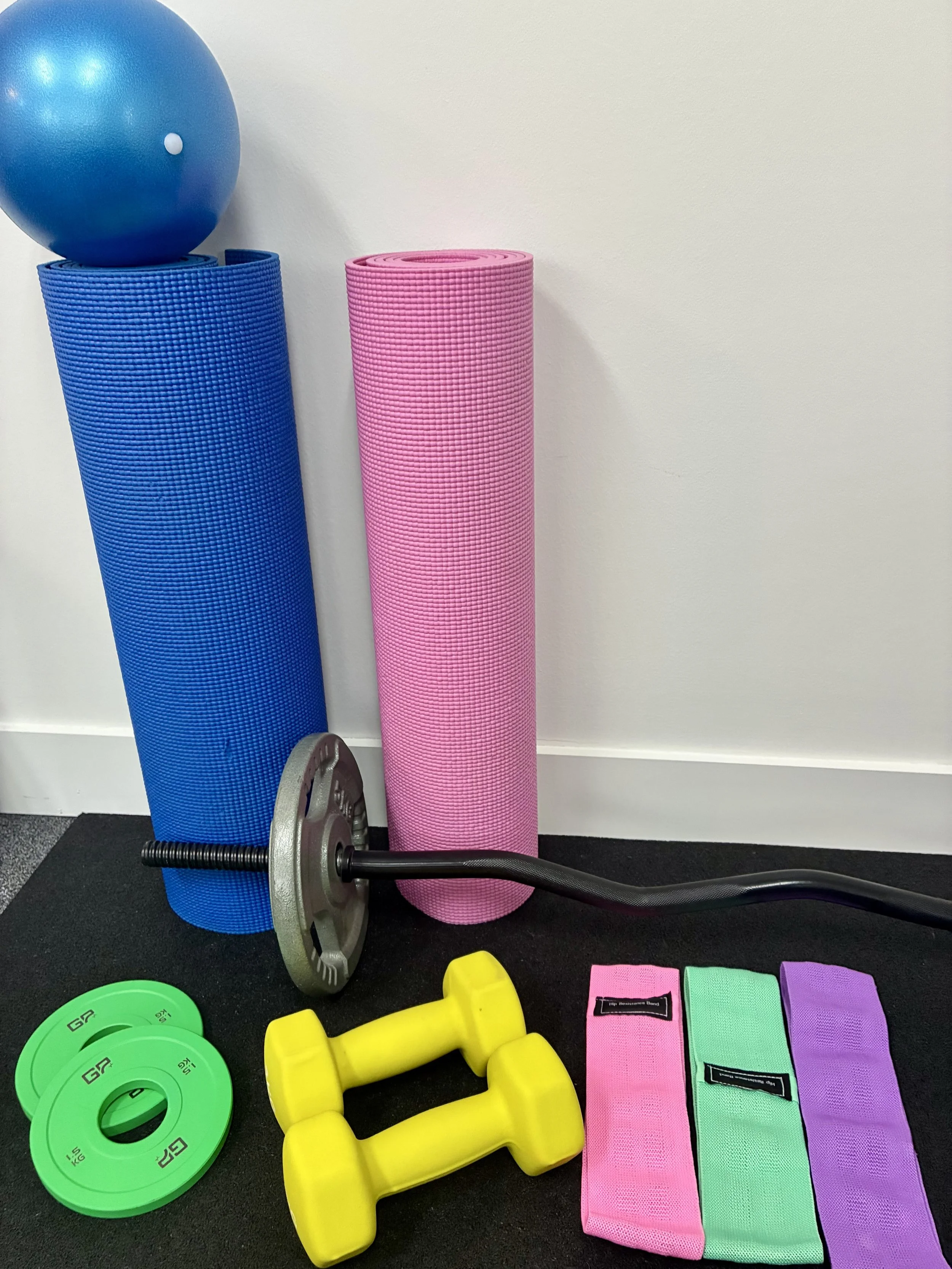 A collection of fitness equipment including a blue exercise ball, a blue yoga mat, a pink yoga mat, a black barbell with weight plates, yellow dumbbells, and three folded resistance bands in pink, green, and purple.