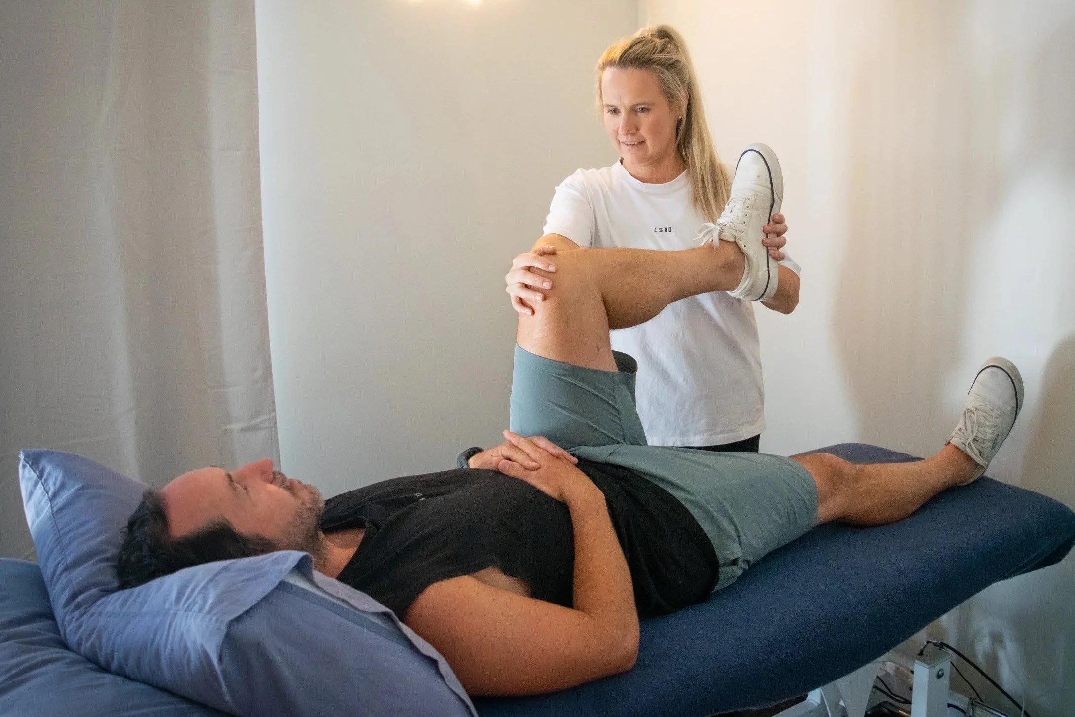 Various kettlebells on the gym floor, including a green 18kg kettlebell and three black kettlebells with different weights, next to a black fan and a piece of gym equipment, against a painted brick wall.