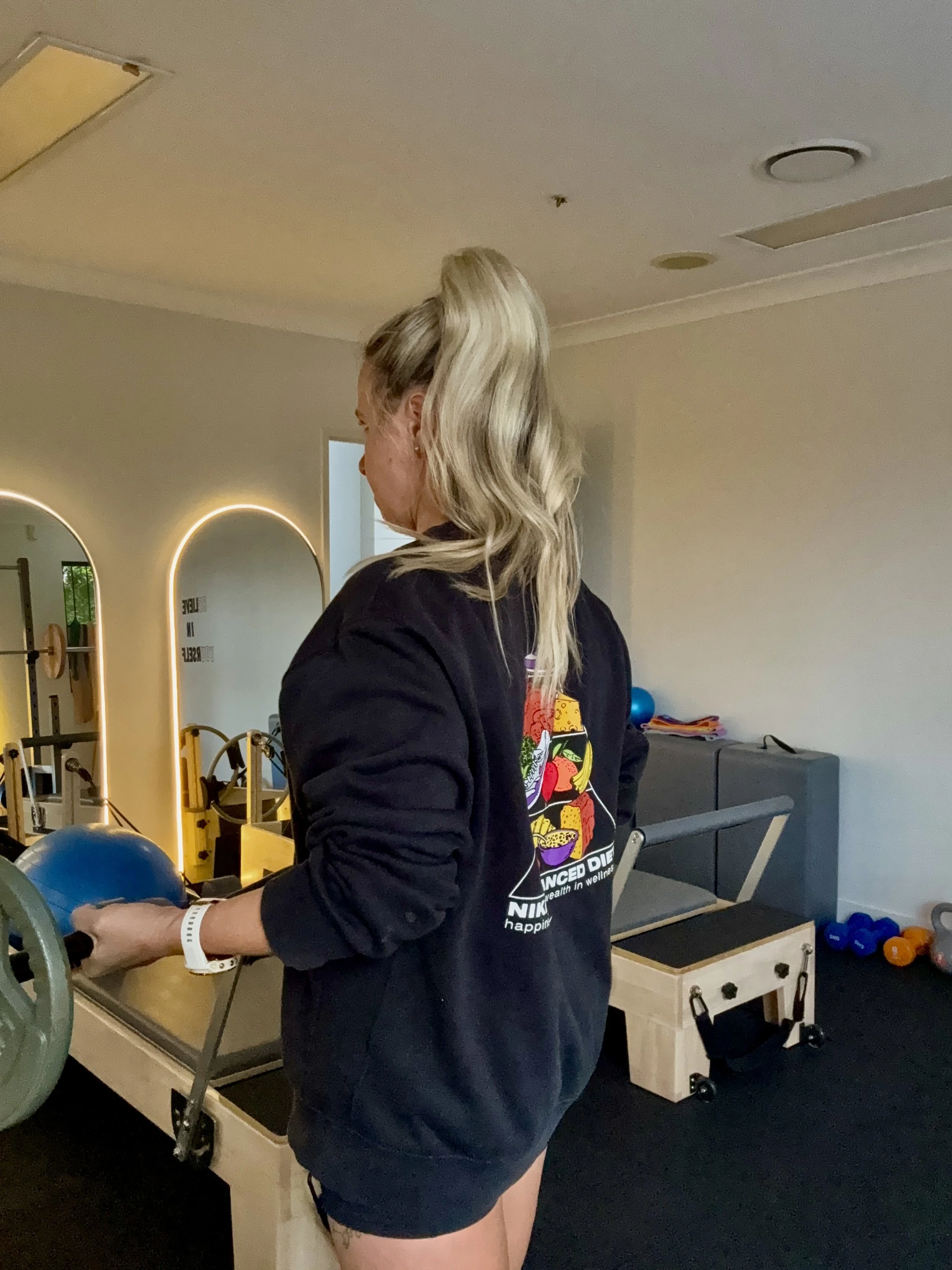 A woman with long, wavy blonde hair wearing a black sweatshirt with colorful fruit graphics, standing in a gym near a workout station with weight plates and other gym equipment.