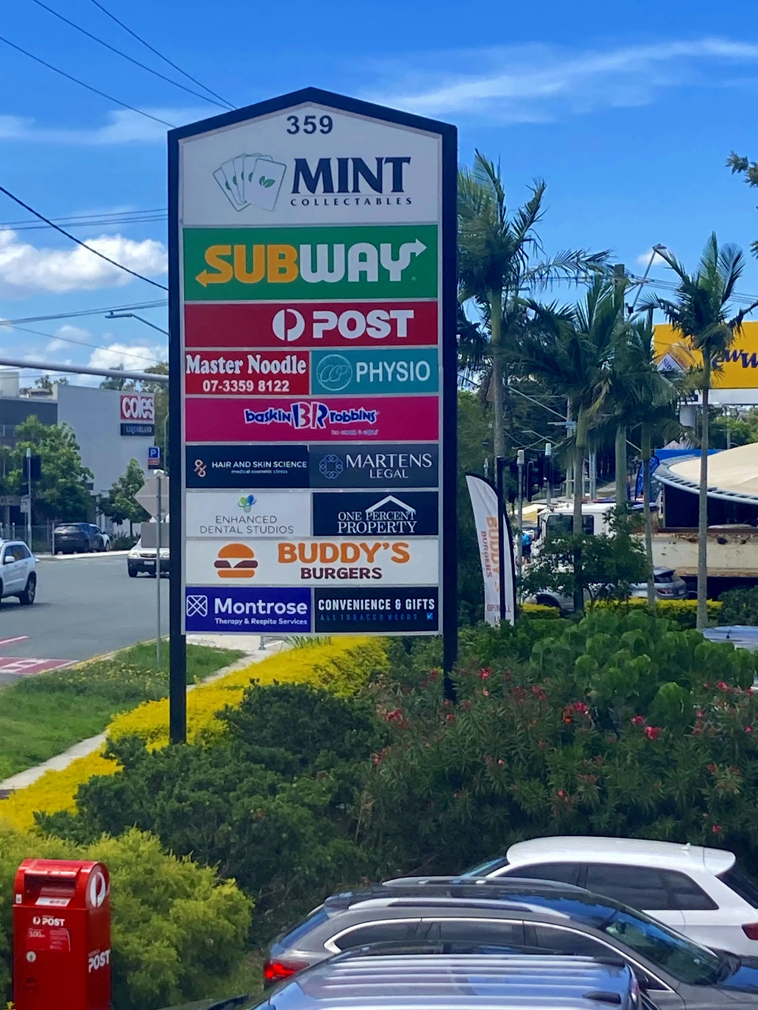 A large vertical sign listing various businesses including Mint Collectables, Subway, Post Office, Master Noodle, Physio, Baskin Robbins, Hair and Skin Science, Martens Legal, Enhanced Dental Studios, One Percent Property, Buddy's Burgers, Montrose Therapy & Respite Services, and Convenience & Gifts. The sign is in a parking lot area with cars and some palm trees visible, against a partly cloudy blue sky.