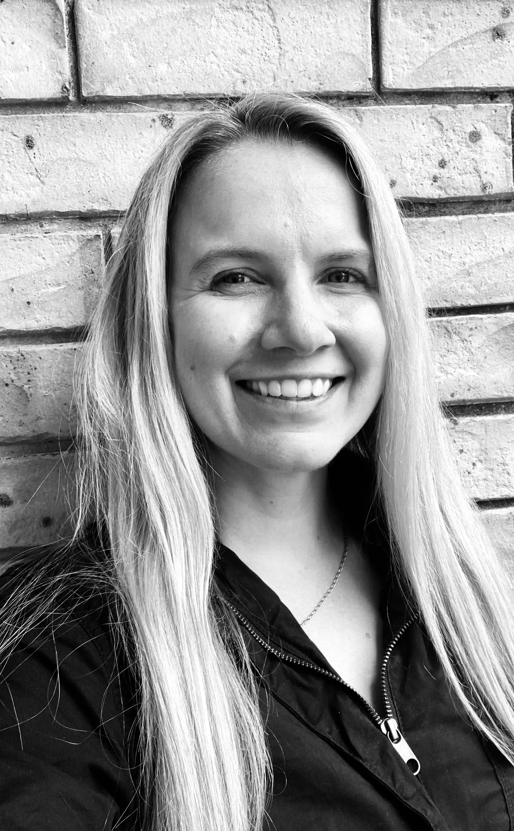 Black and white photo of a woman with long blonde hair smiling, standing against a brick wall.