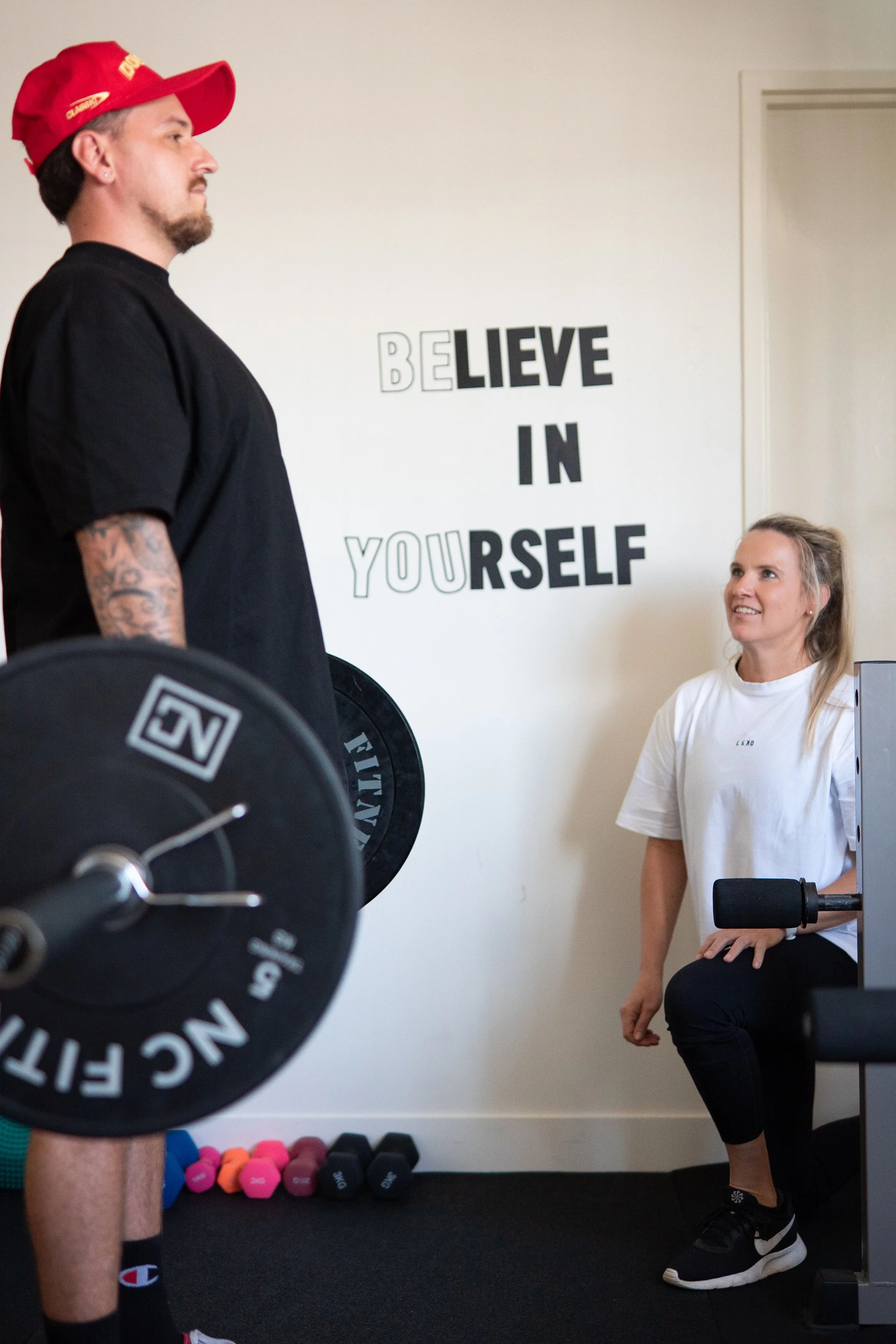 A man holding a weight in a gym standing in front of a seated woman who is smiling and looking at him, with a motivational quote on the wall that reads 'BELIEVE IN YOURSELF'.