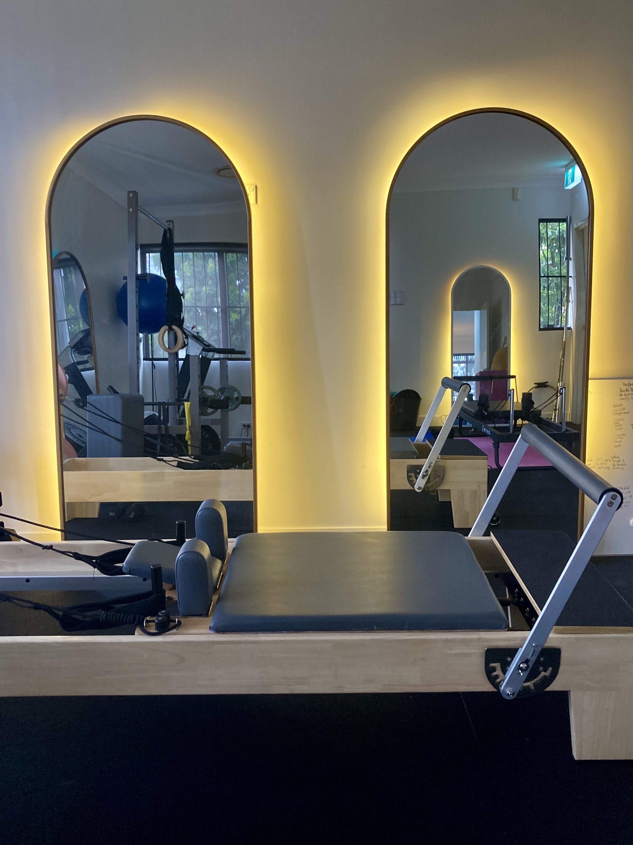 Studio with Pilates reformer, resistance bands, and gym equipment reflected in arched mirrors with yellow backlighting.
