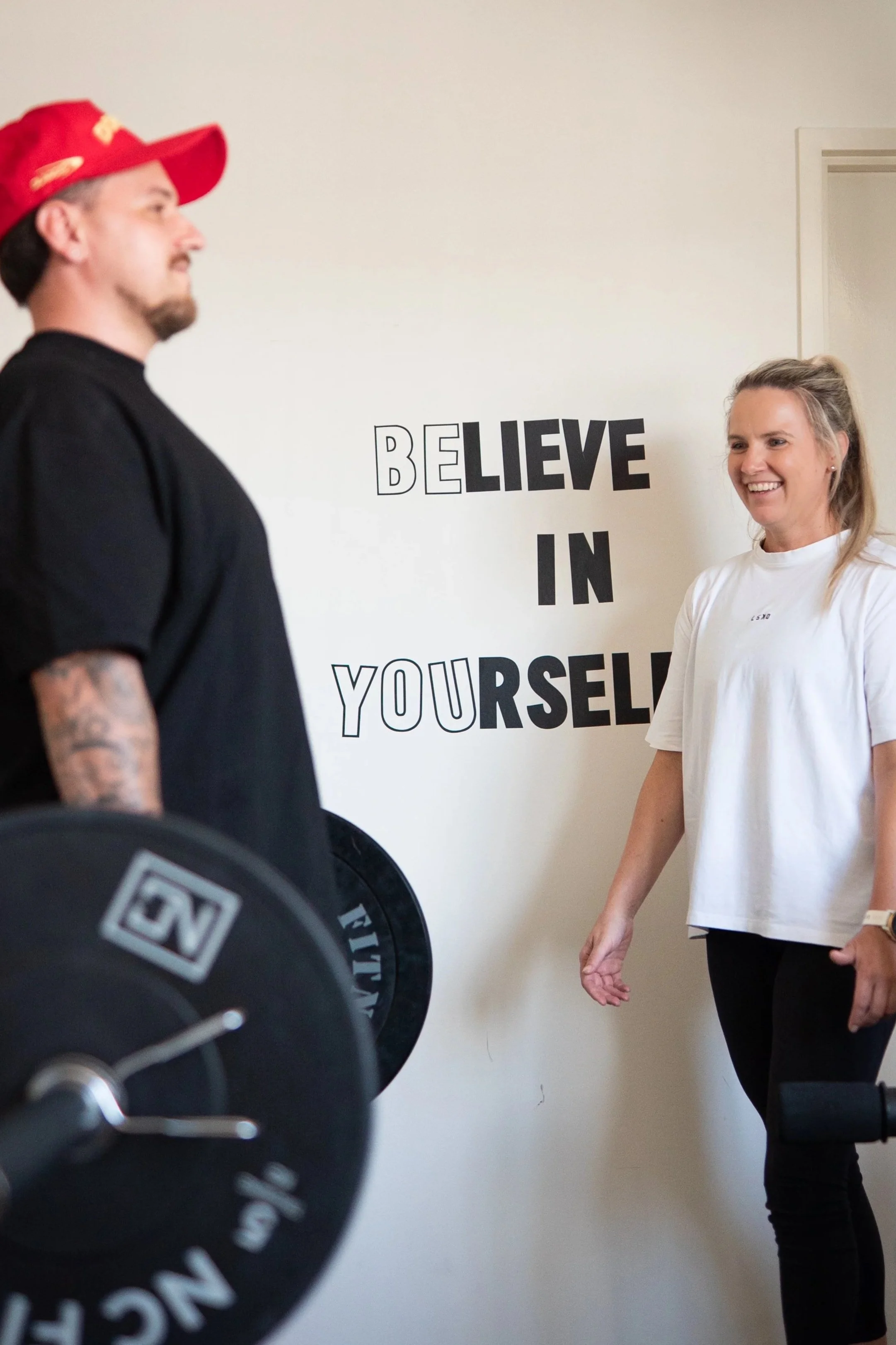 A woman smiling and standing in front of a motivational wall quote that says "BELIEVE IN YOURSELF," while a man in a red cap and black shirt faces her during a workout session in a gym.