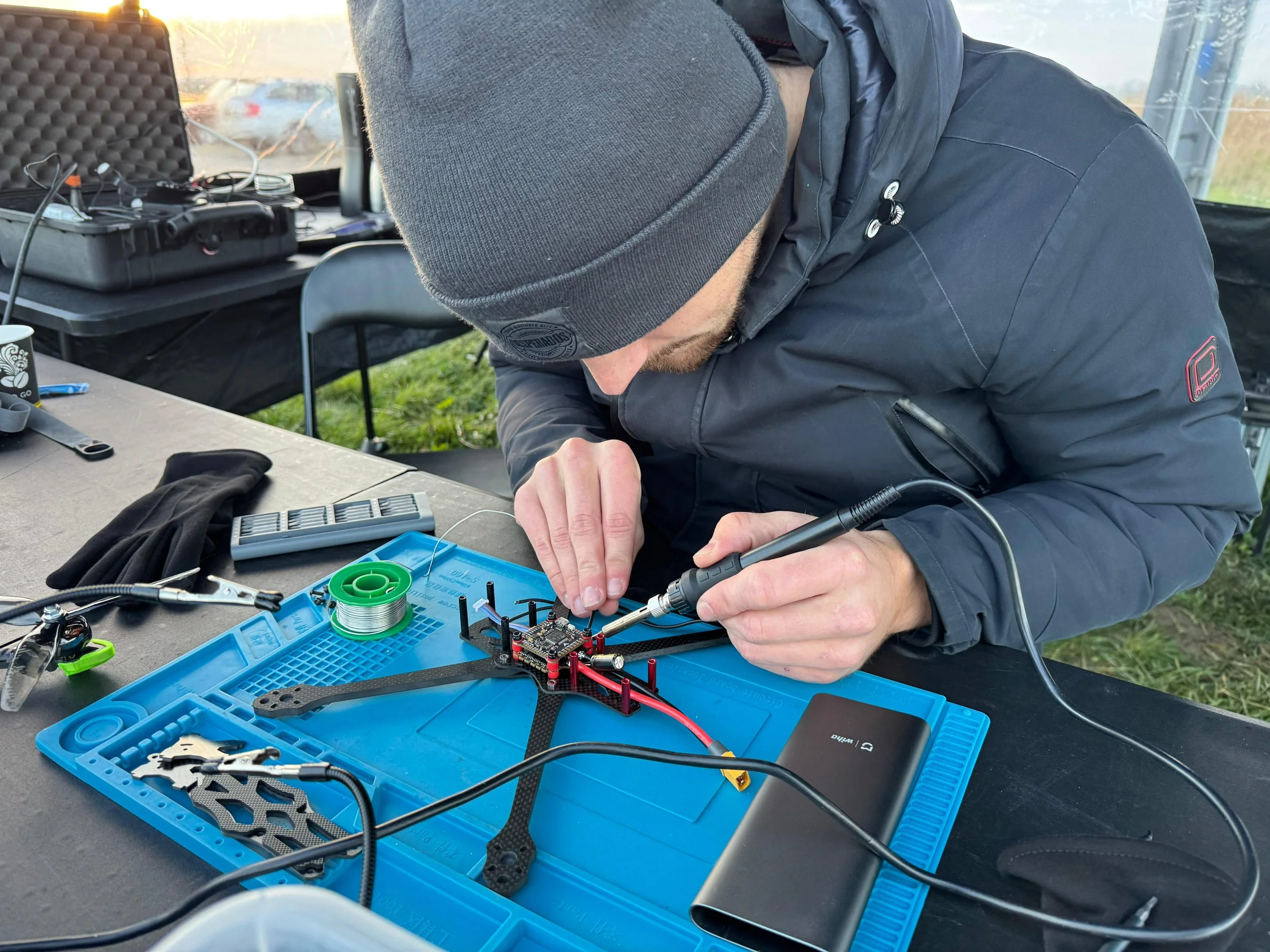 Person wearing a gray beanie and black jacket soldering electronic components on a drone frame in an outdoor workspace.