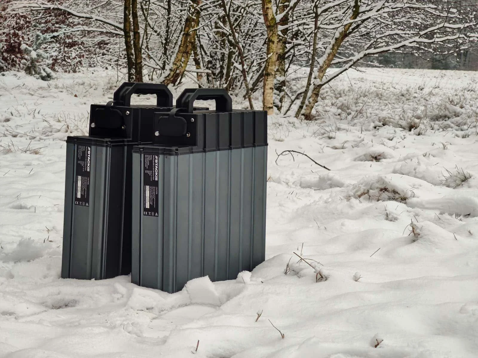 Four metal battery packs placed on snowy ground near leafless trees in winter landscape.