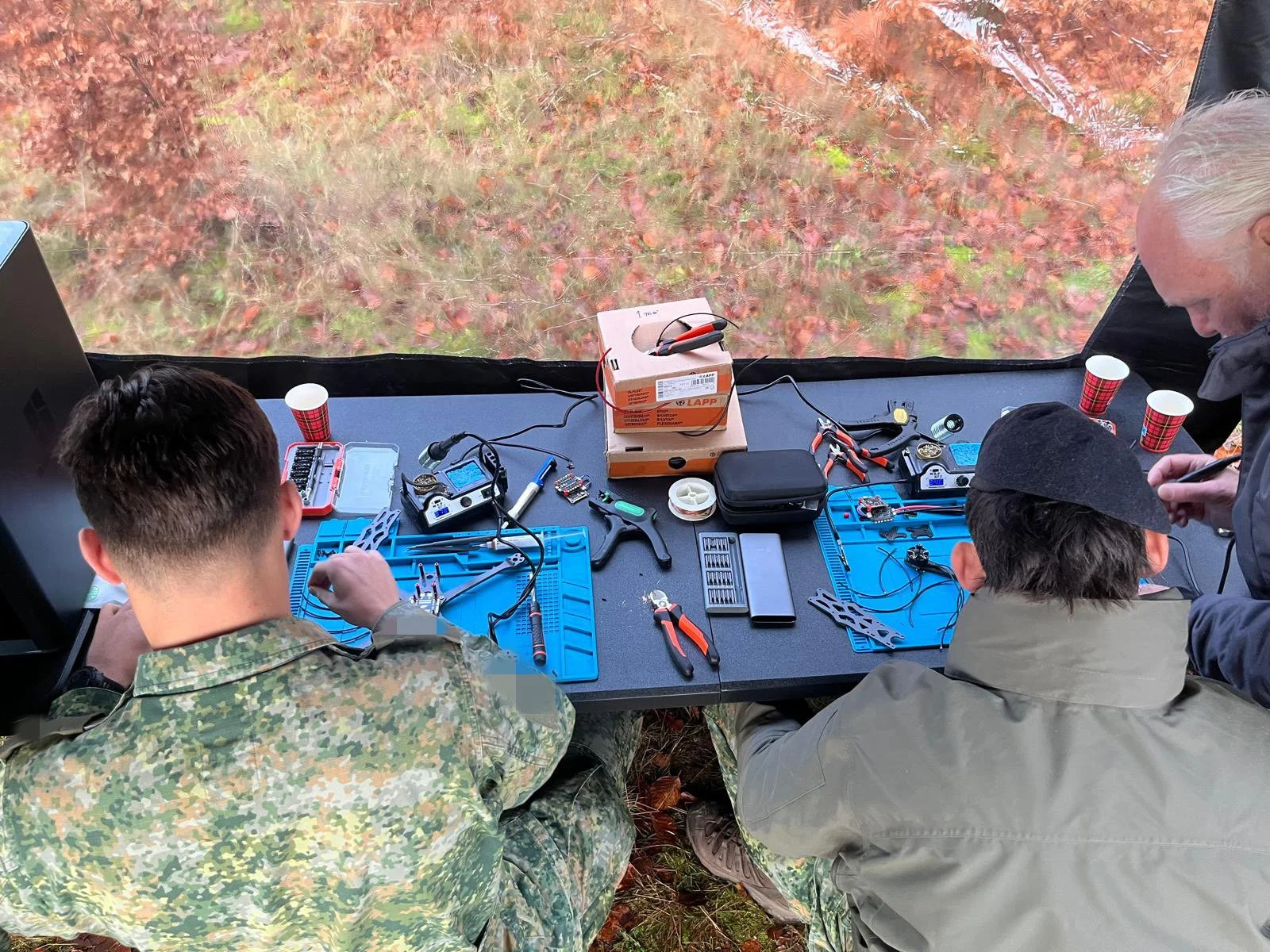 Three people sit at a table outdoors working on electronic devices and tools, with a view of autumn leaves outside.