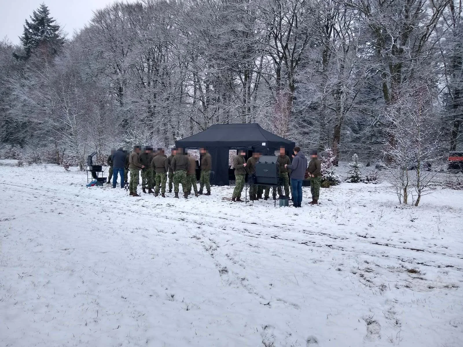 A group of people, some in military uniforms, gathered under a black canopy tent in a snowy field surrounded by trees, possibly filming or conducting an outdoor activity.