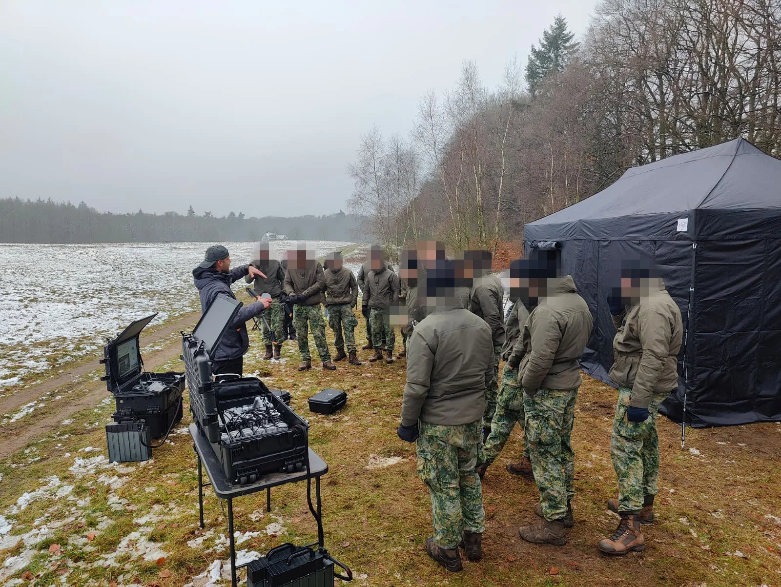 Group of military personnel listening to instructor near a lake, outdoors with no snow on the ground, some snow patches, and a tent.