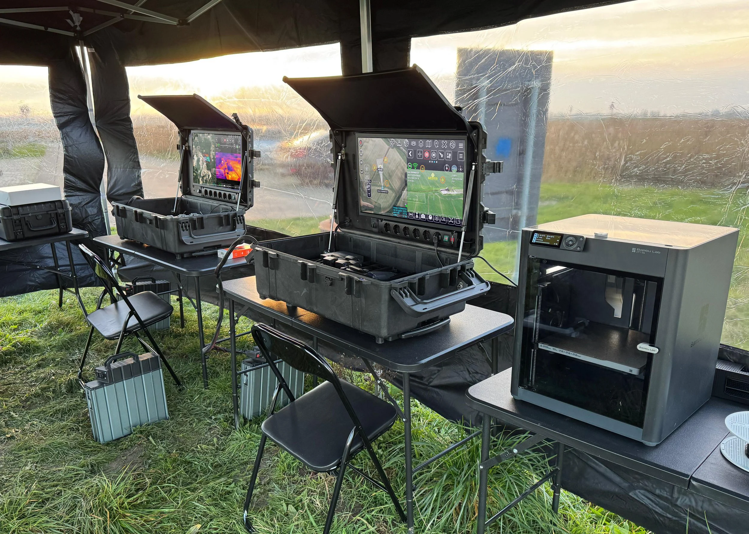 Field testing setup with monitors, laptops, and equipment inside a tent with plastic walls, set on grass outdoors during sunset.