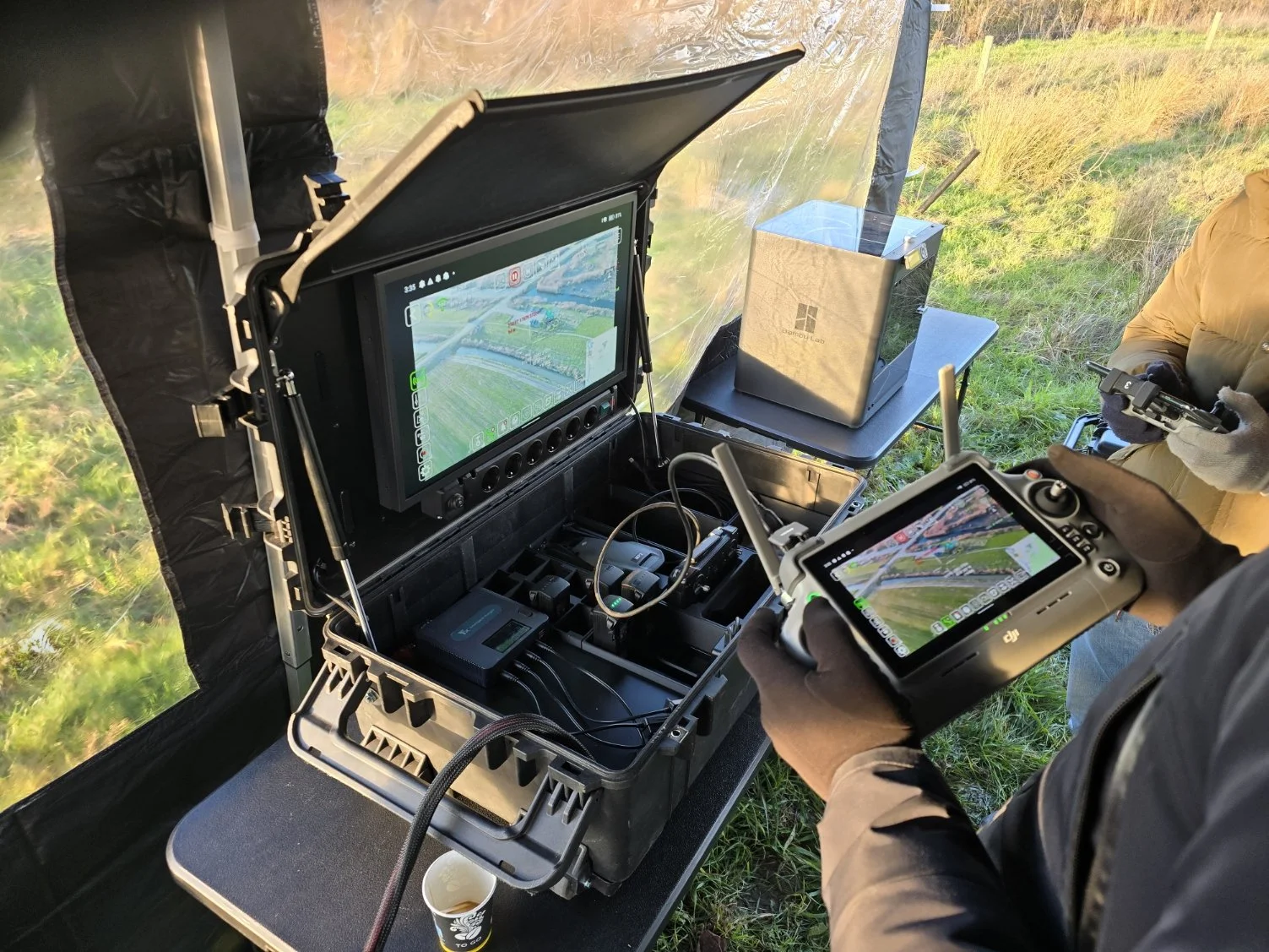 Two people operating a drone control station outdoors, with a monitor and remote controllers, surrounded by grassy field.
