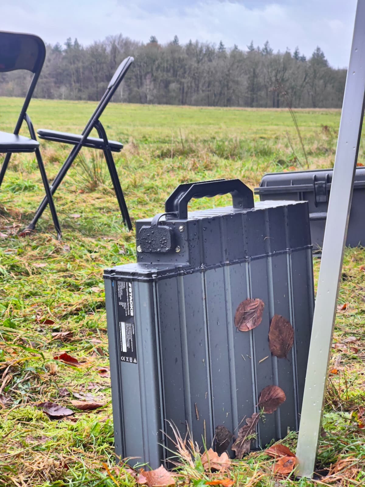 A black portable battery pack with leaves on its surface, placed outdoors on grass, with two folding chairs and a bag in the background, and a forested area with trees in the distance.