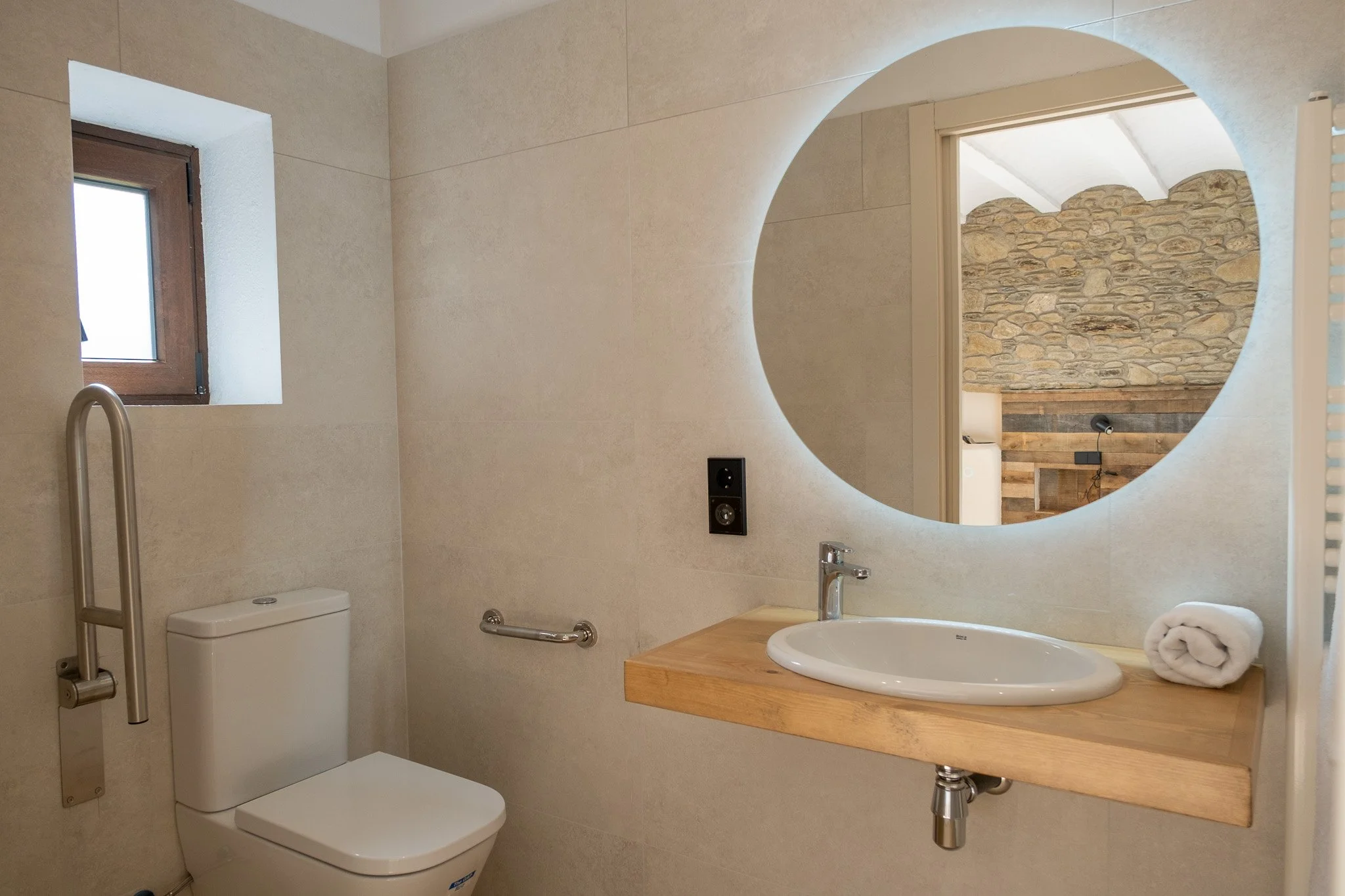 Baño con inodoro, lavabo con lavabo de madera, espejo redondo con iluminación y pared de piedra visible en reflejo.