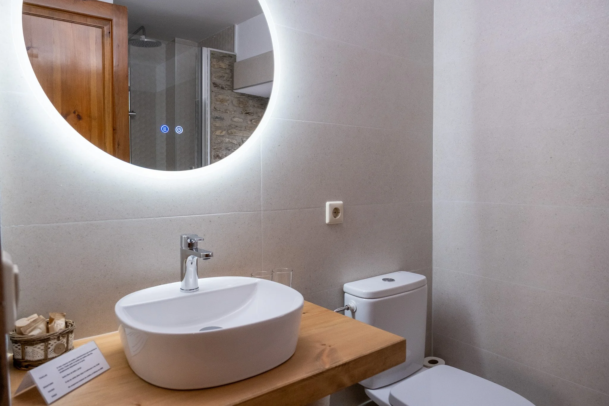 Baño con lavabo de cerámica sobre un mostrador de madera, espejo redondo con luz, inodoro y paredes revestidas en tono claro, con una toma de corriente en la pared.