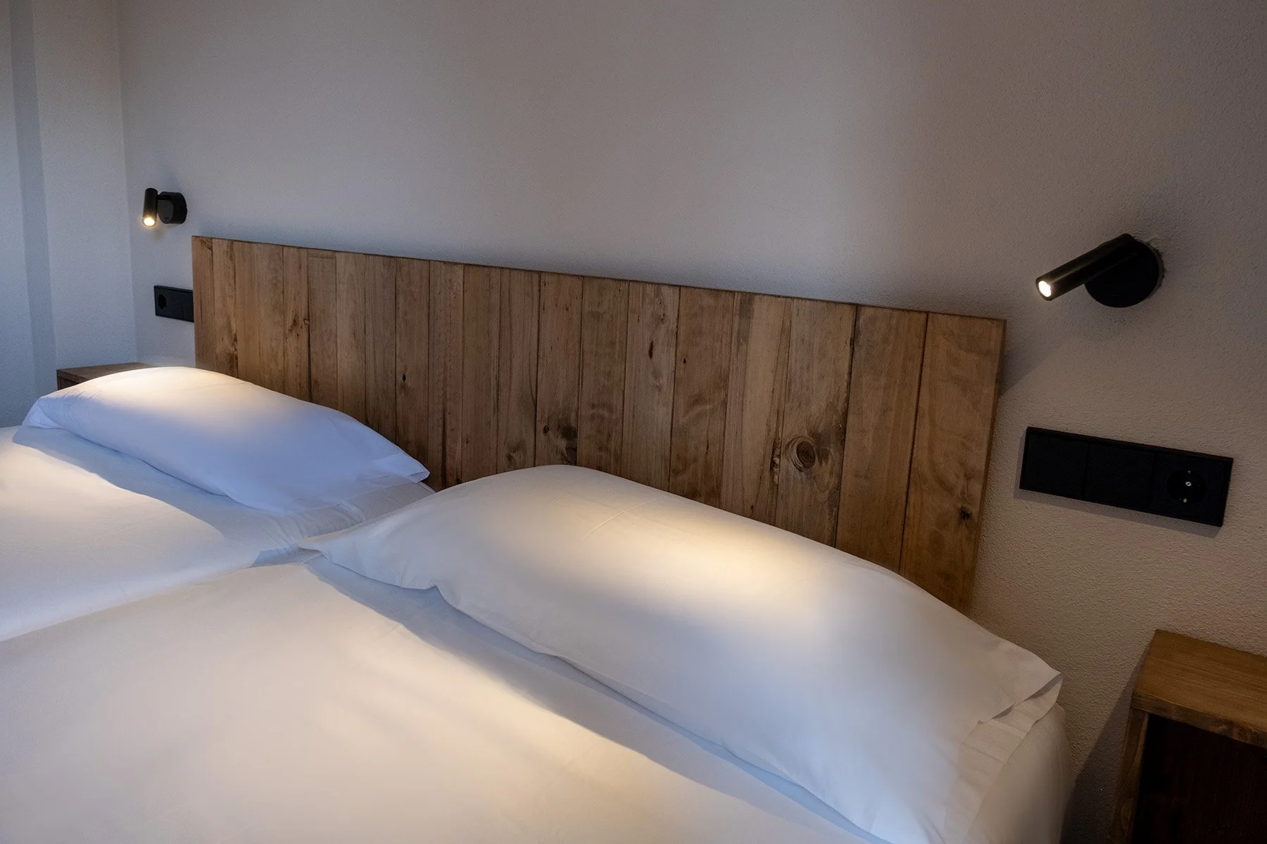 Cama con cabecera de madera en habitación moderna con paredes blancas y lámparas negras ajustadas en la pared.