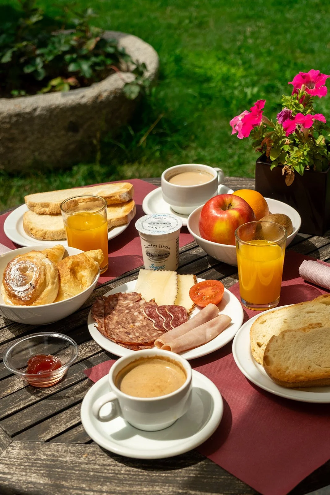 Desayuno al aire libre con pan, croissants, jugo, café, yogur, fruta y embutidos en una mesa de madera con plantas y césped en el fondo.